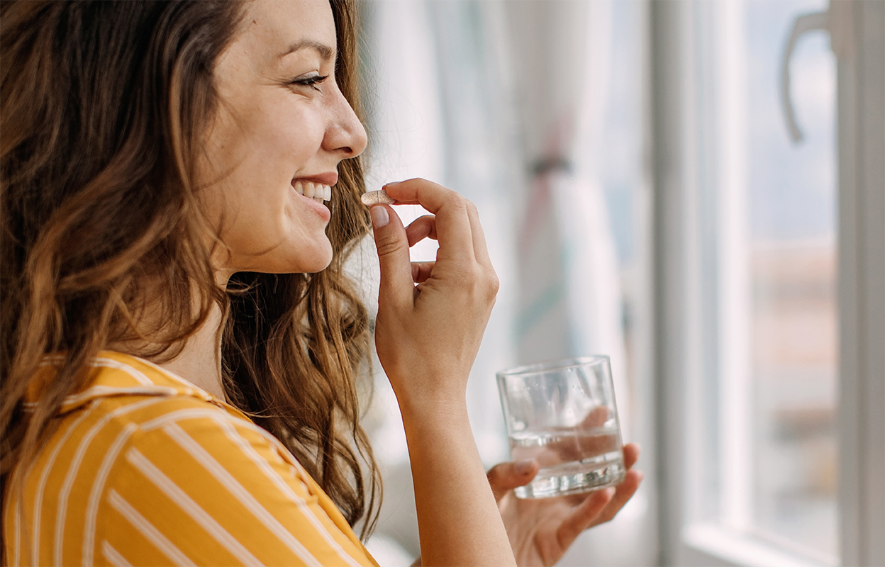 Smiling woman in a striped yellow dress taking a supplement with a glass of water near a bright window.