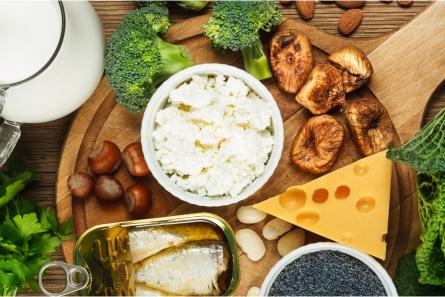A selection of calcium-rich foods including milk, broccoli, cheese, sardines, figs, and nuts, artfully arranged on a wooden board.