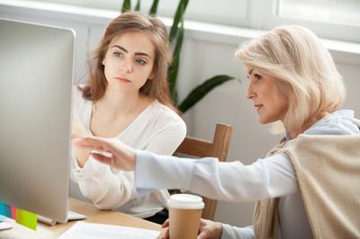 Senior and young female colleagues discuss online project look at pc screen, older mentor teaches young woman explains corporate software work, aged executive helps intern, teamwork on computer task