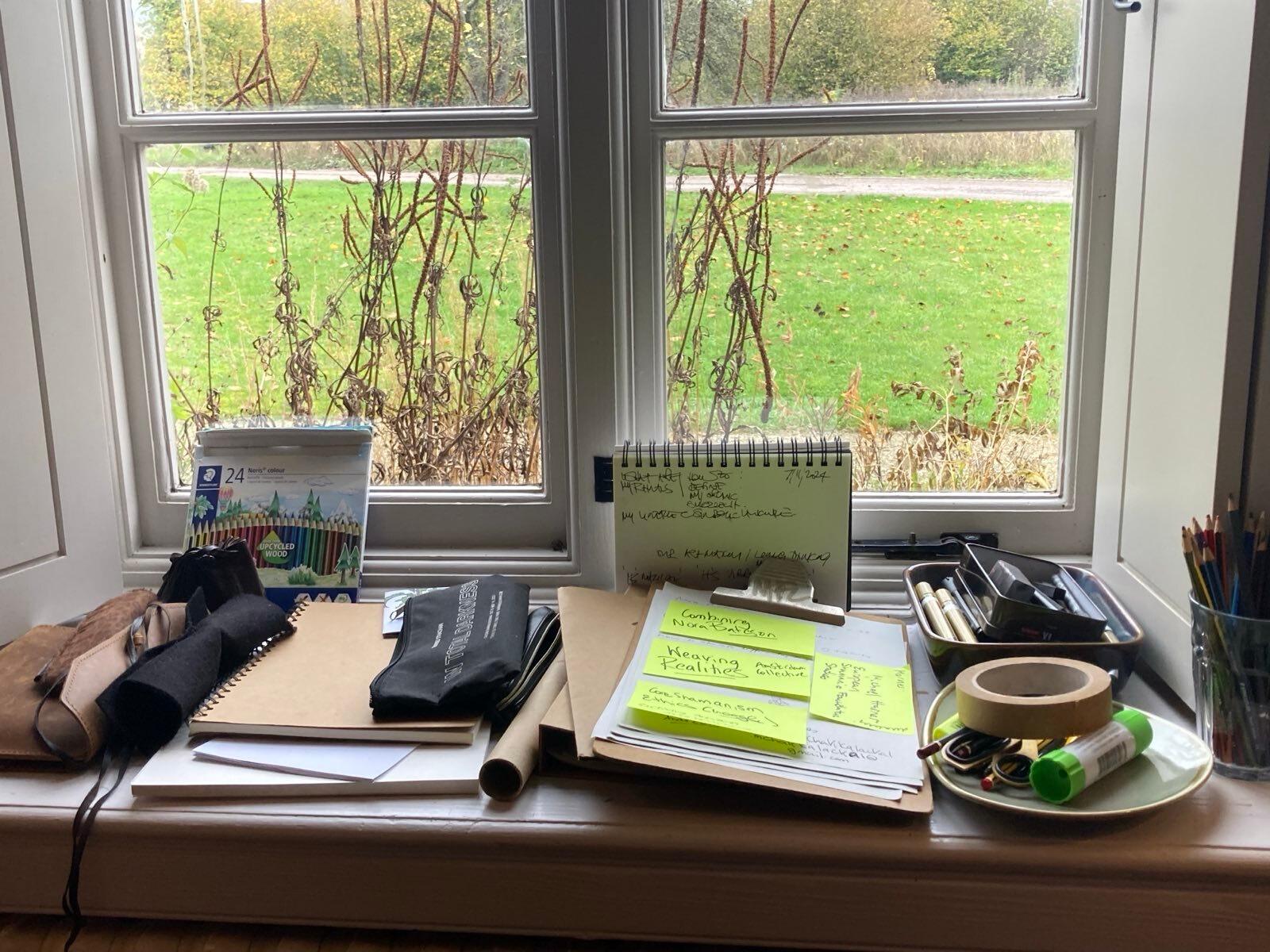 A writing desk with paper and pens, and a large window behind it looking out into a garden.