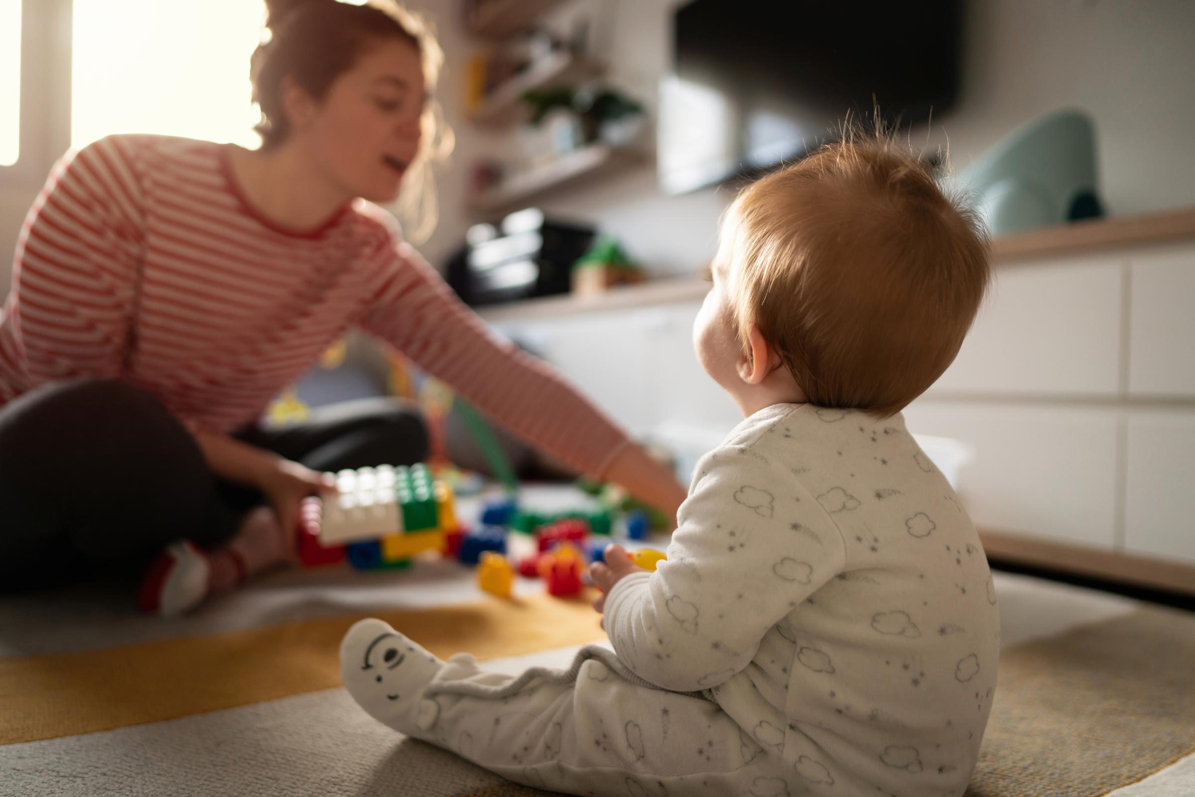 Back view of a Caucasian baby boy sitting on the floor, having fun with toys and looking at his mother.