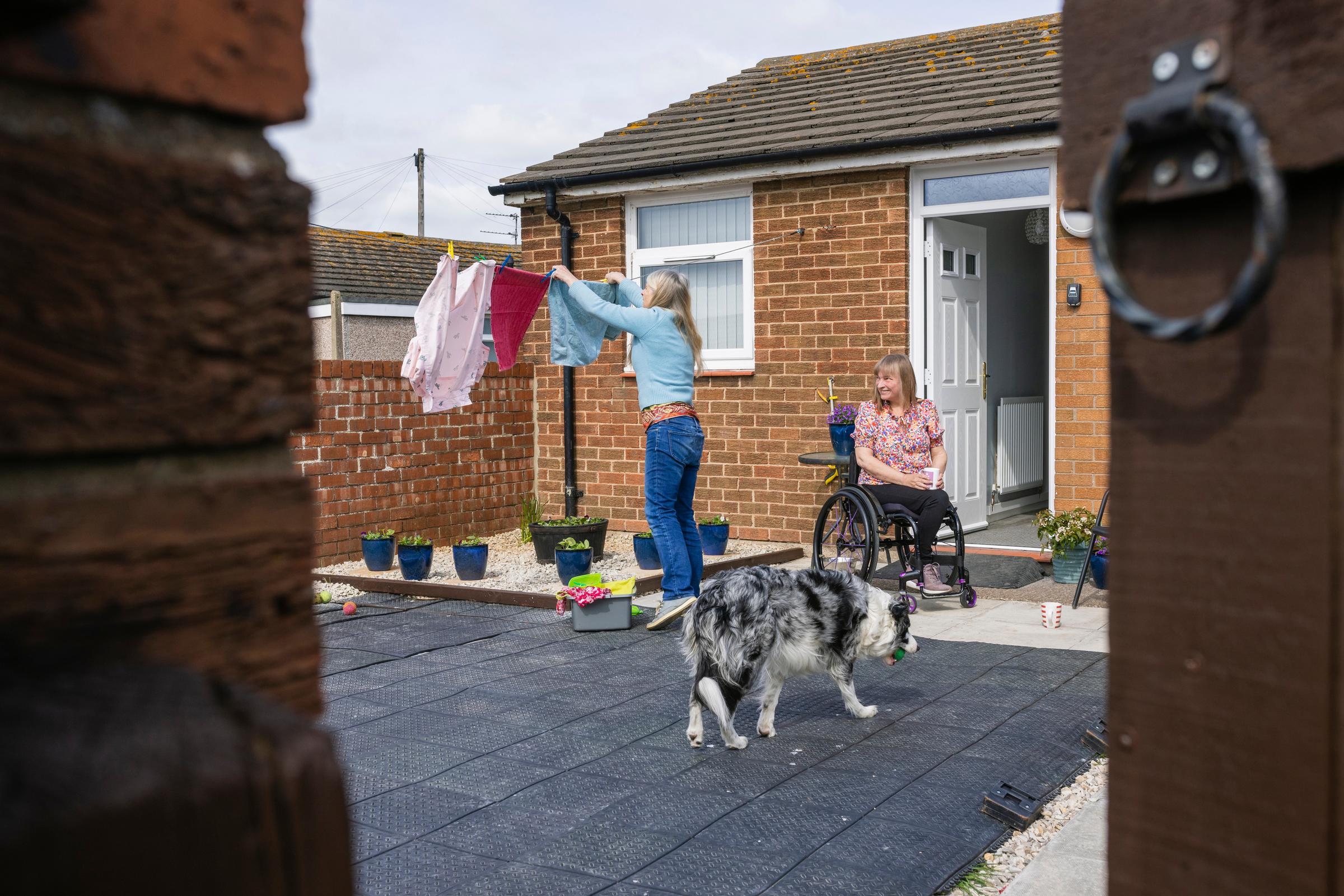 A wide angle view of a woman and her friend who is a wheelchair user having a catch up. She is helping her friend hang out her washing on a clear dry day outside of her home in Amble in the North East of England