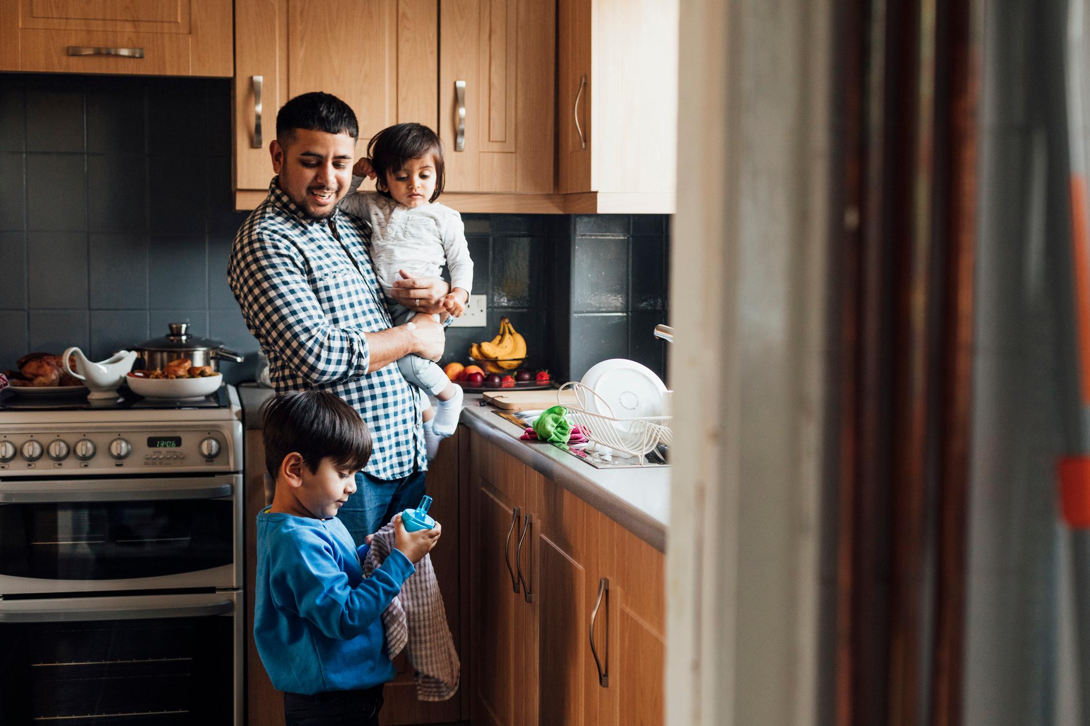 A father with his sons at home in the kitchen together. He is carrying his youngest son while looking at his eldest son, who is drying a reusable bottle with a dishtowel.