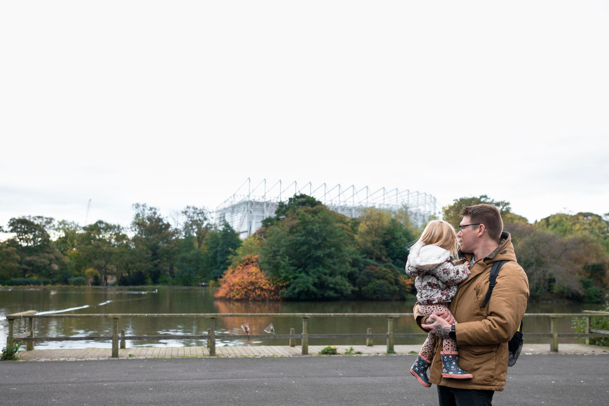 Father and his young daughter walking alongside a lake in a public park. They are looking at ducks  floating on the lake.