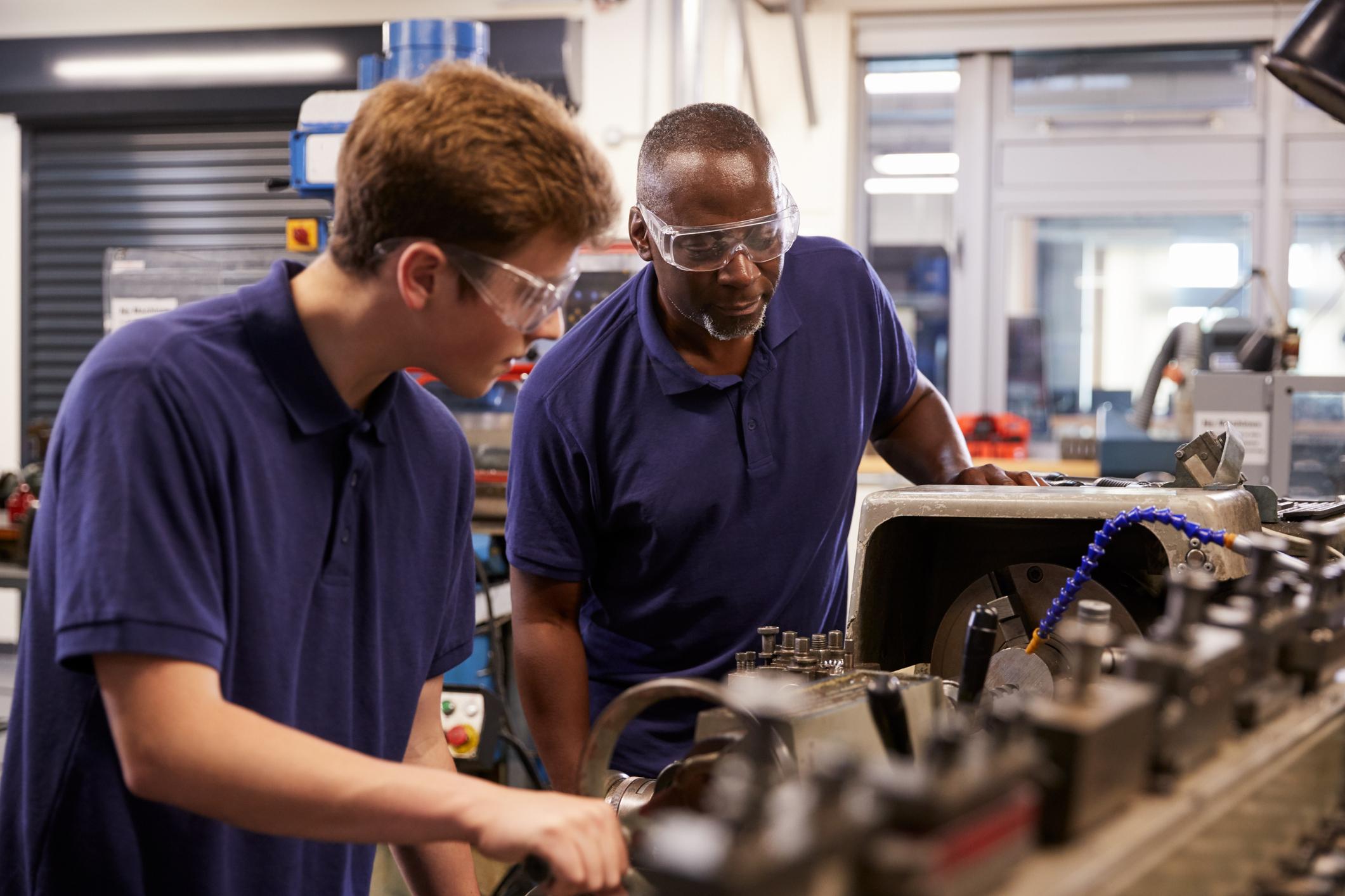 Engineer supervising a teenage apprentice using a lathe.