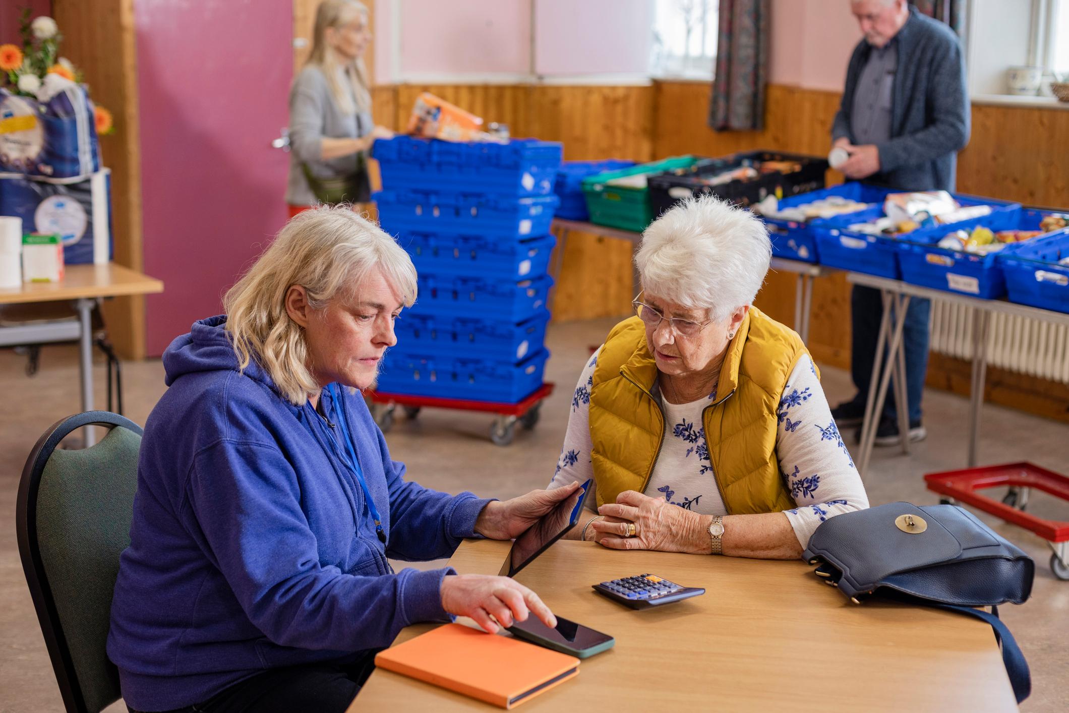 Two older women at a community church where a food bank is being run by volunteers in the North East of England. They are sitting at a table together, using a calculator to work out budgets, helping each other to save money during the inflation.