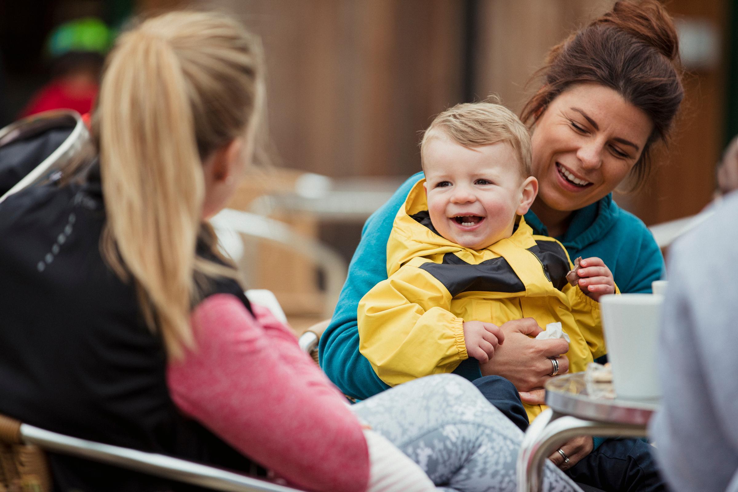 Woman at cafe table with a child on her lap talking to another woman and smiling.