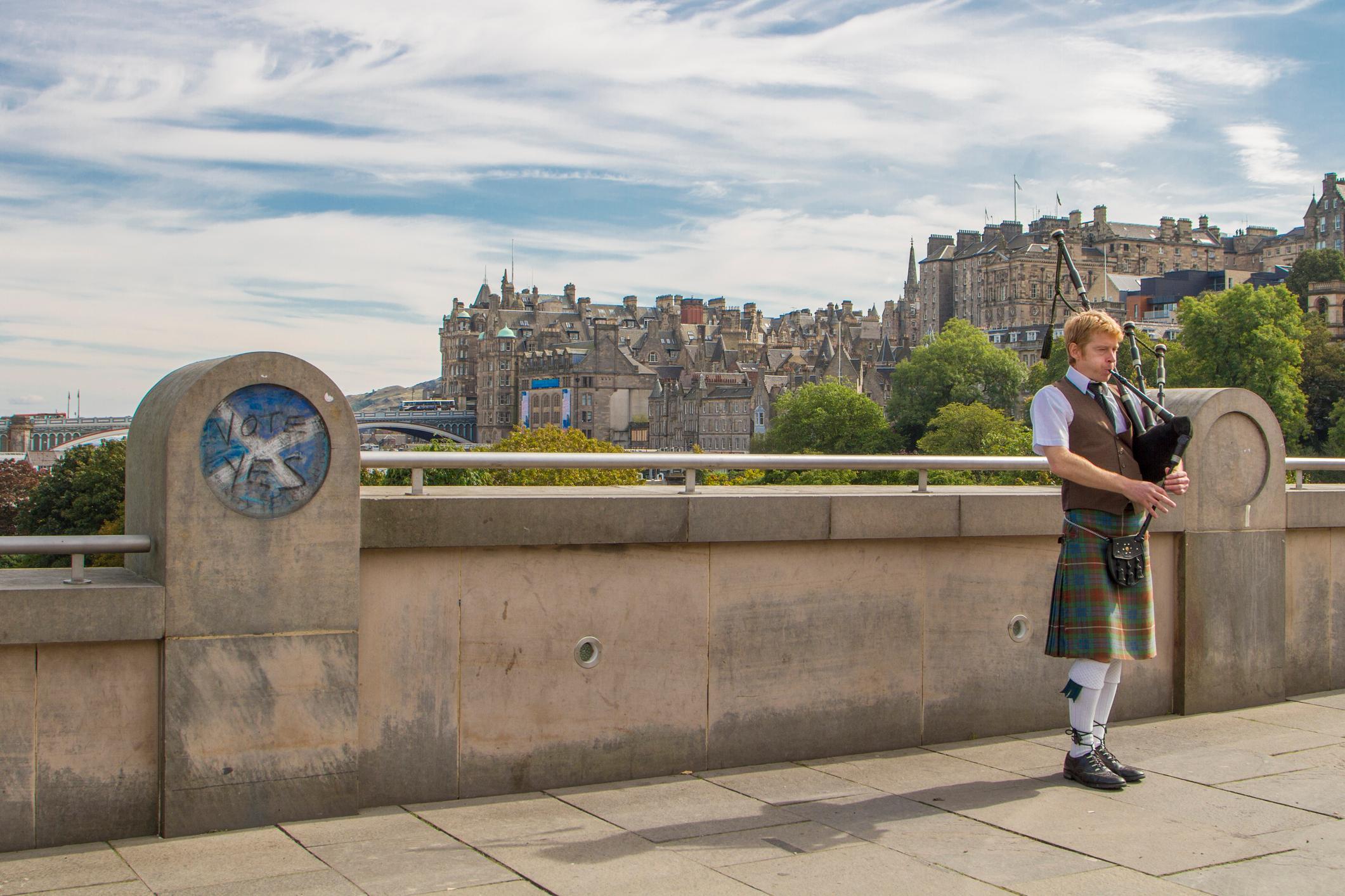 Scottish man in traditional costume playing bagpipes with a graffiti painted 'Vote Yes' sign on wall next to him.