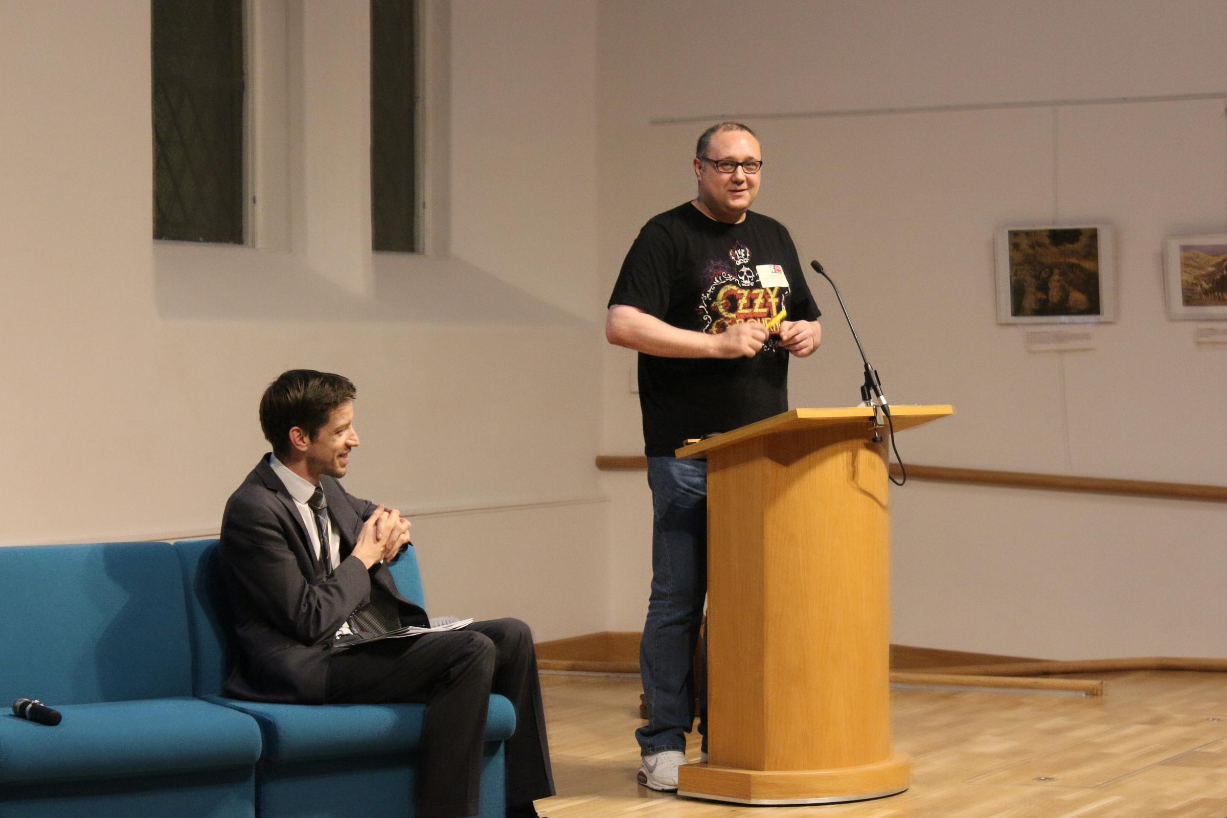 A photograph showing Tony Gibson, former co-chair of DFLP, giving a speech. John Alexander, former co-chair of DFLP and former leader of Dundee City Council, is sitting down and smiling. The photograph was taken by Faith in Community Dundee.