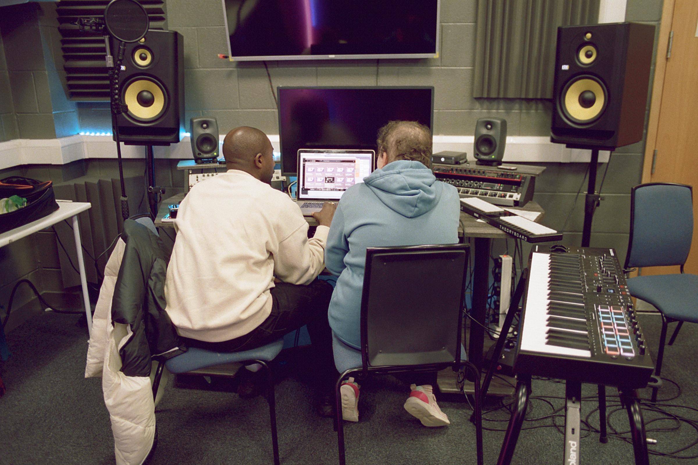 Two people sat, backs to camera, facing a laptop and other music editing equipment in a recording studio