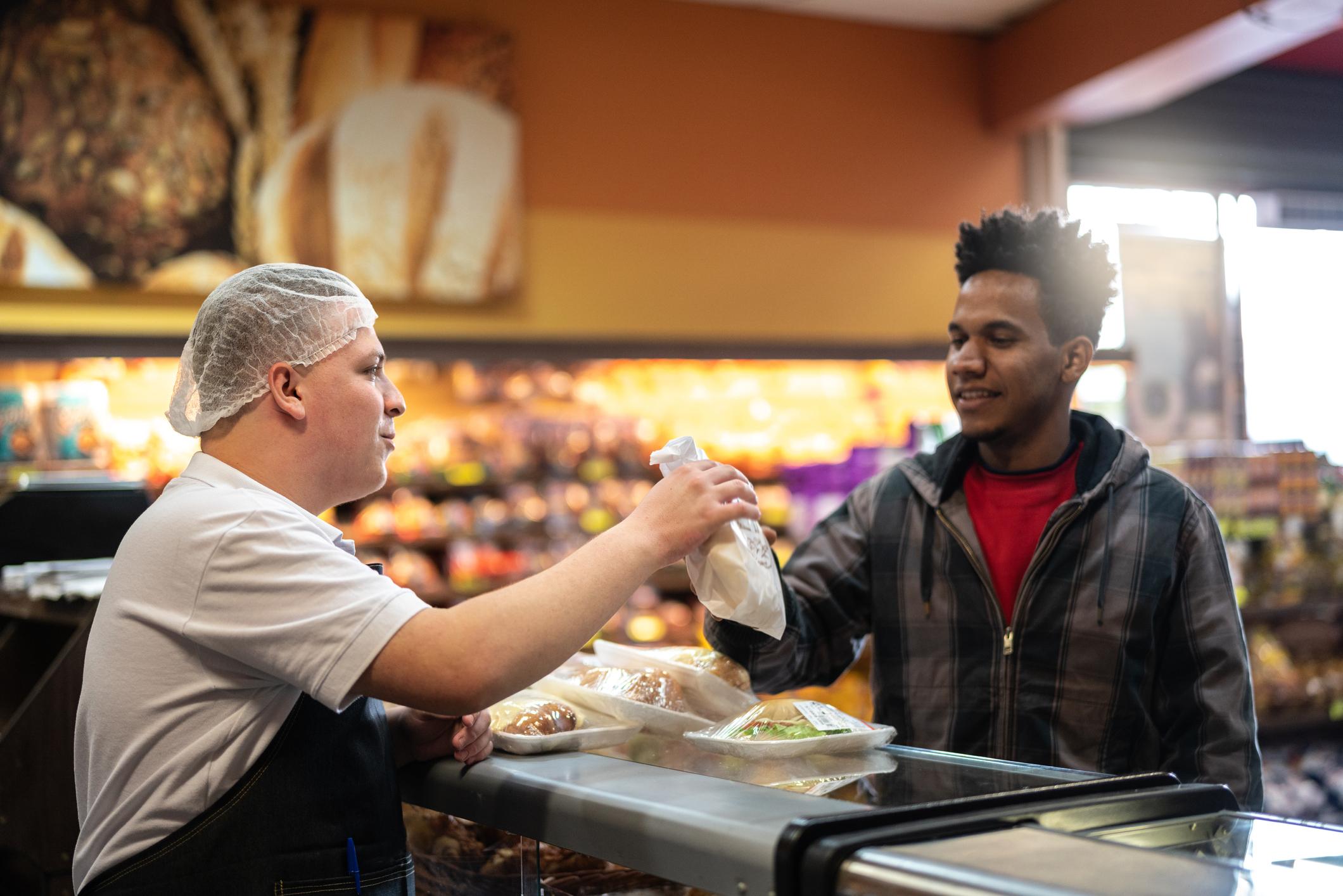 A customer is buying a sandwhich from a shop assistant, they are smiling.