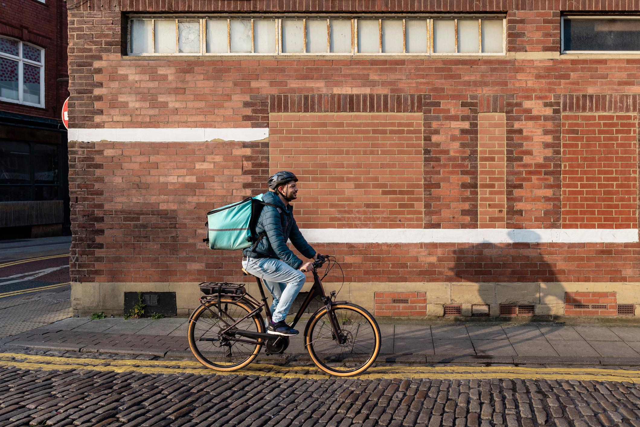 A delivery man wearing a bicycle helmet and food delivery bag, riding his bike on a cobbled road.