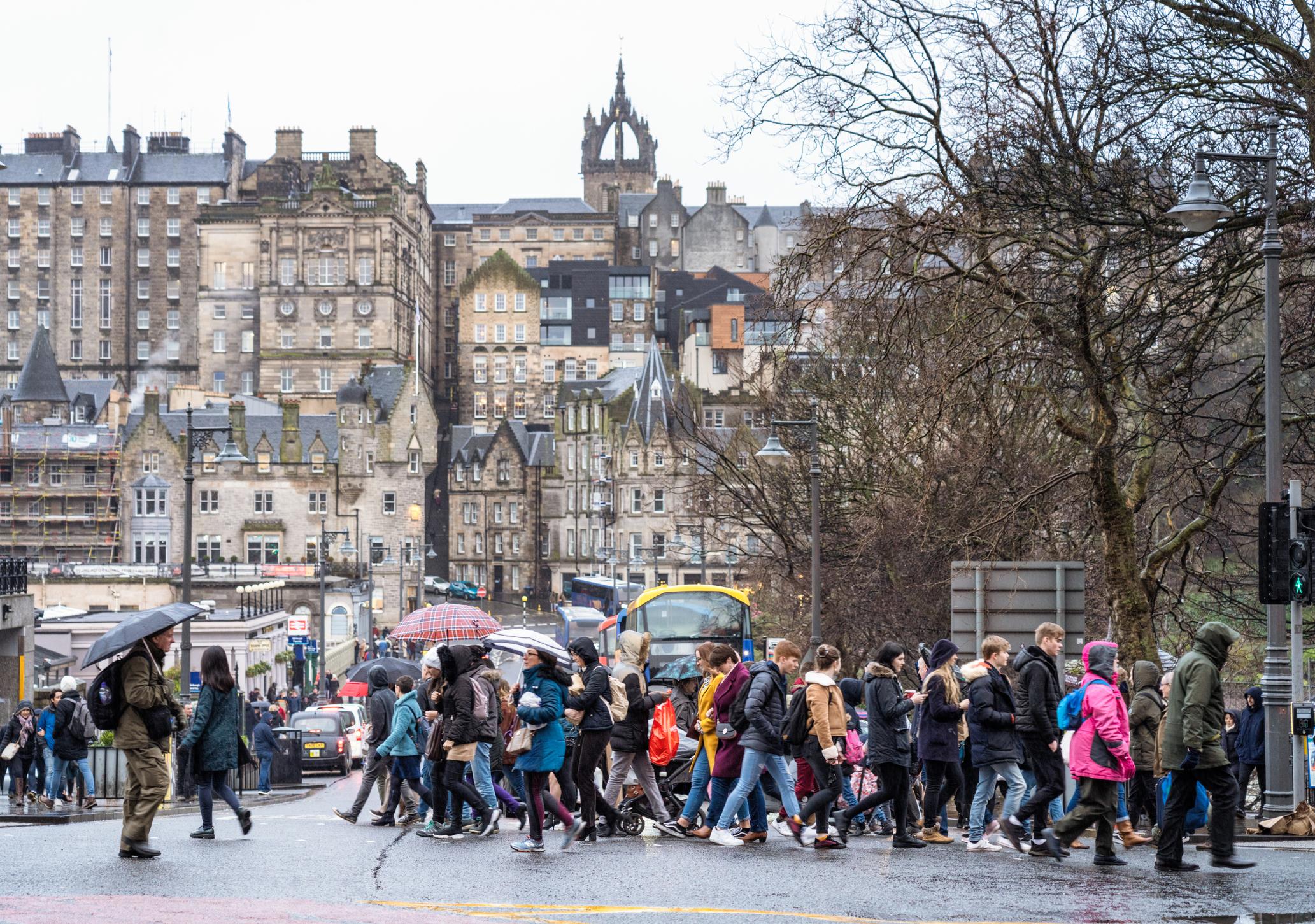 Pedestrians crossing Waverley Bridge in central Edinburgh, with a skyline of the city's Old Town in the background.