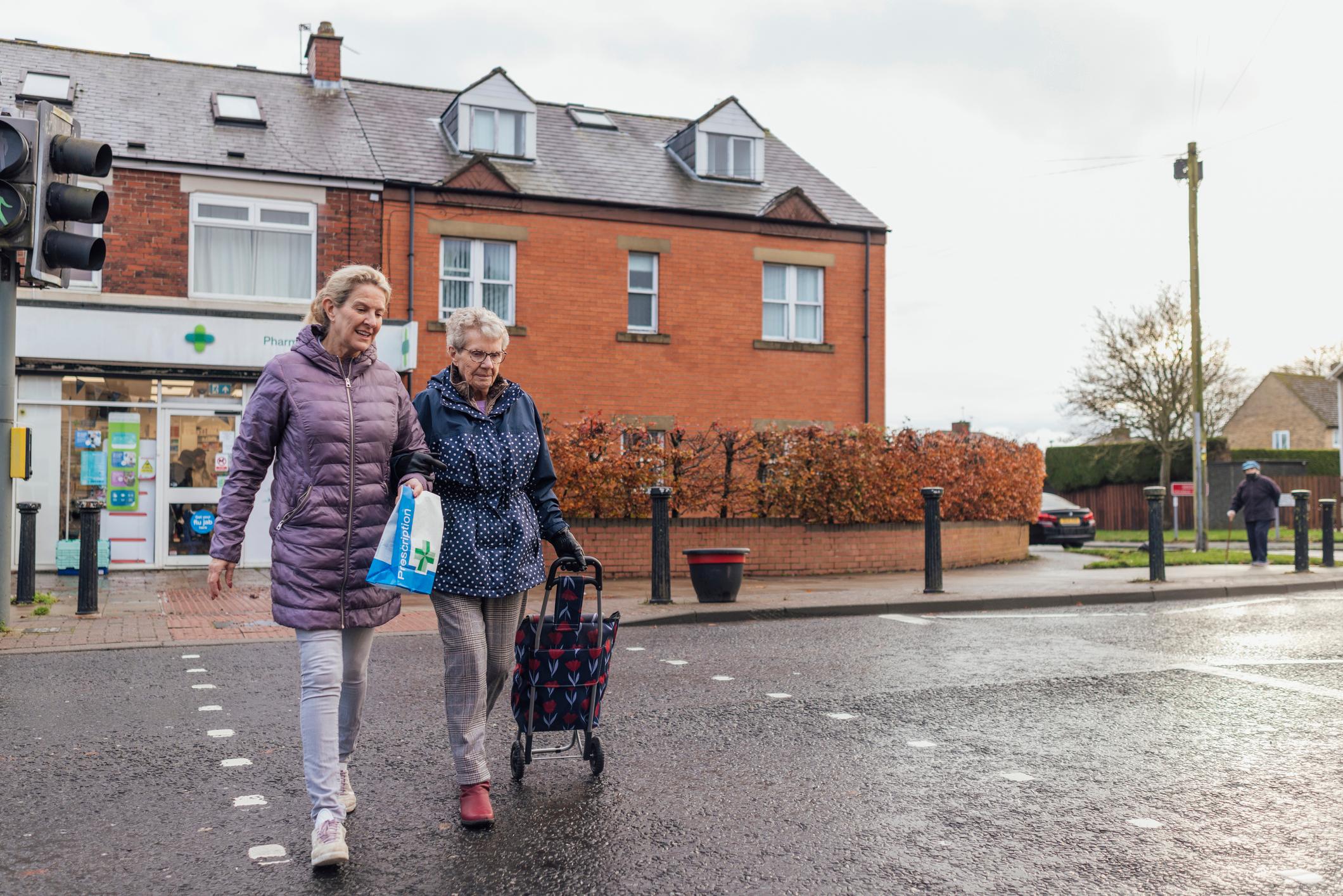 A woman walking arm in arm with an older woman for support and assistance as they cross a road. They have just picked up the senior woman's prescription at the pharmacy behind them.