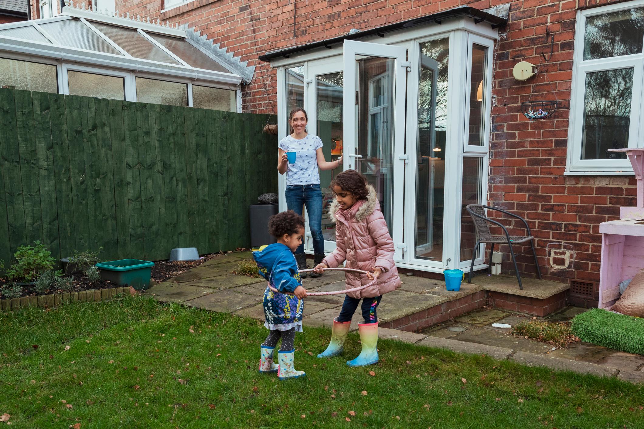 Two young girl siblings playing in the garden together, their mother is watching them while having a hot drink.