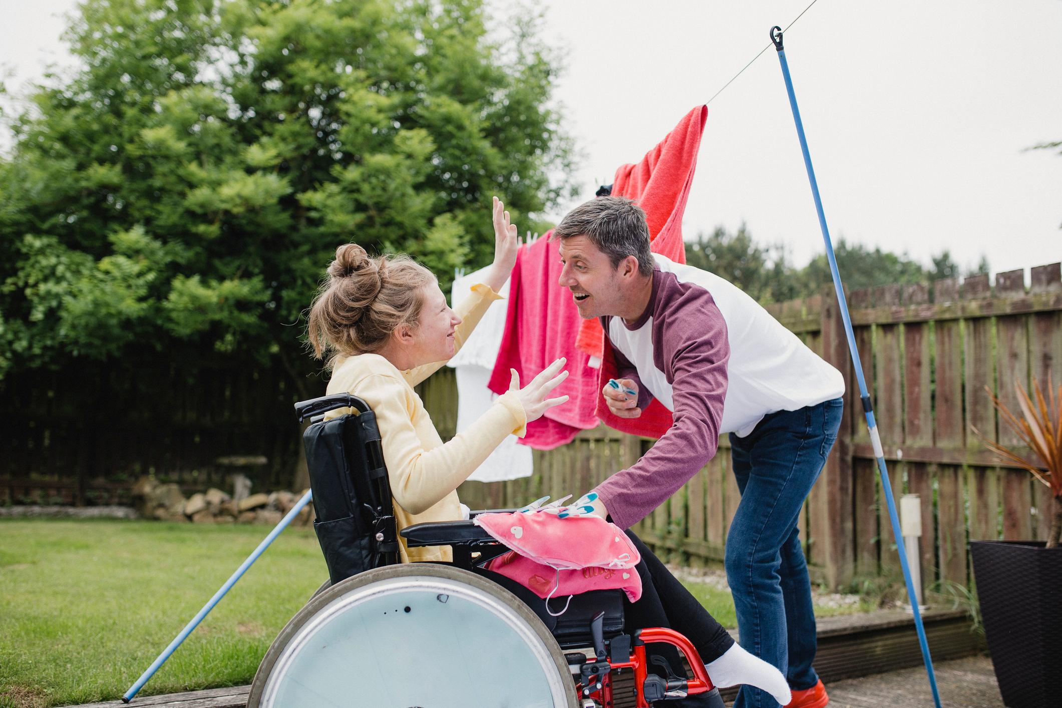 Disabled teenager is in her wheelchair in the garden while her father hangs clothes on the washing line.