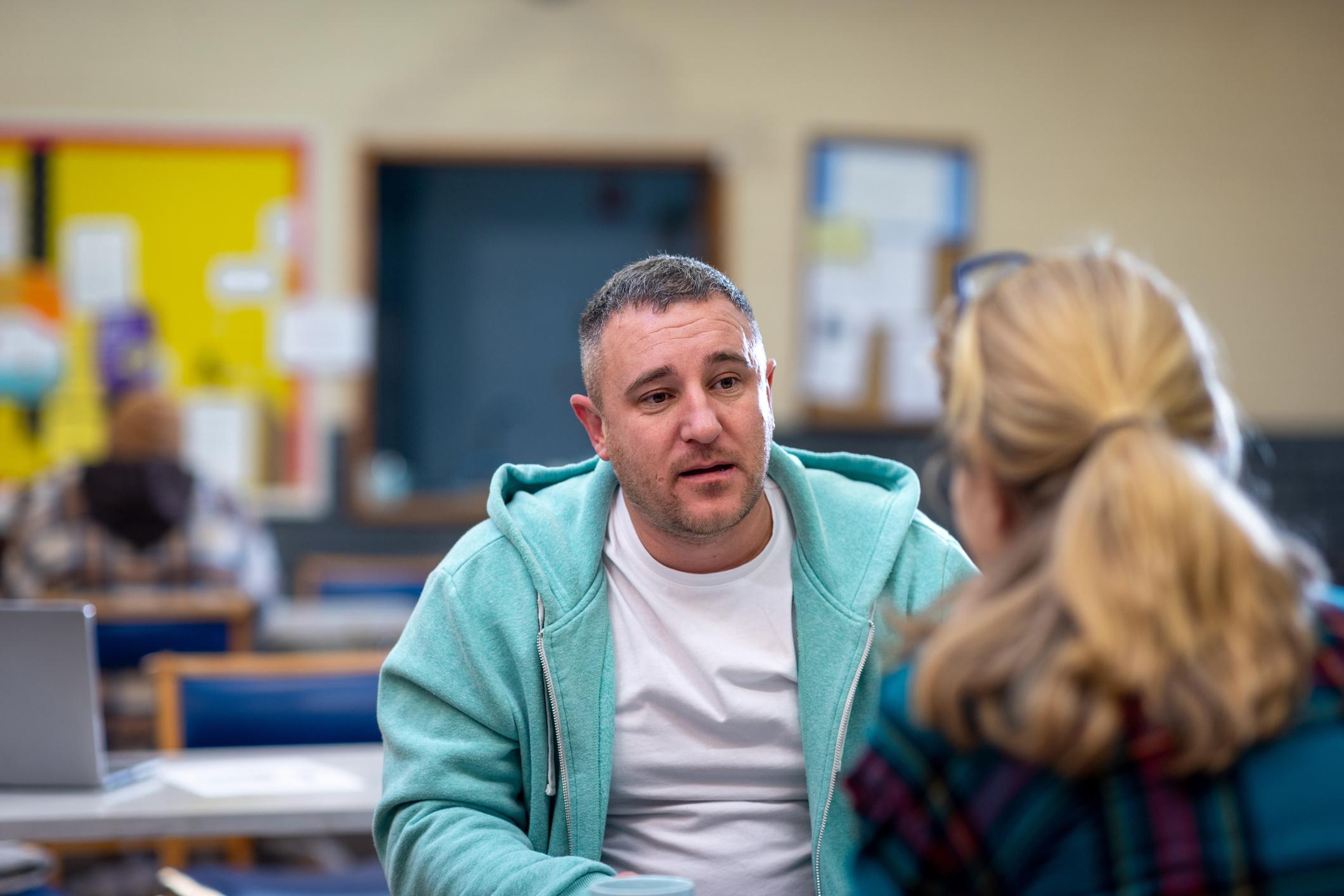 Over-the-shoulder shot of a male adult having a one-to-one with another adult about their mental well-being. The community centre is located in Seaton Deleval in the North East of England.
