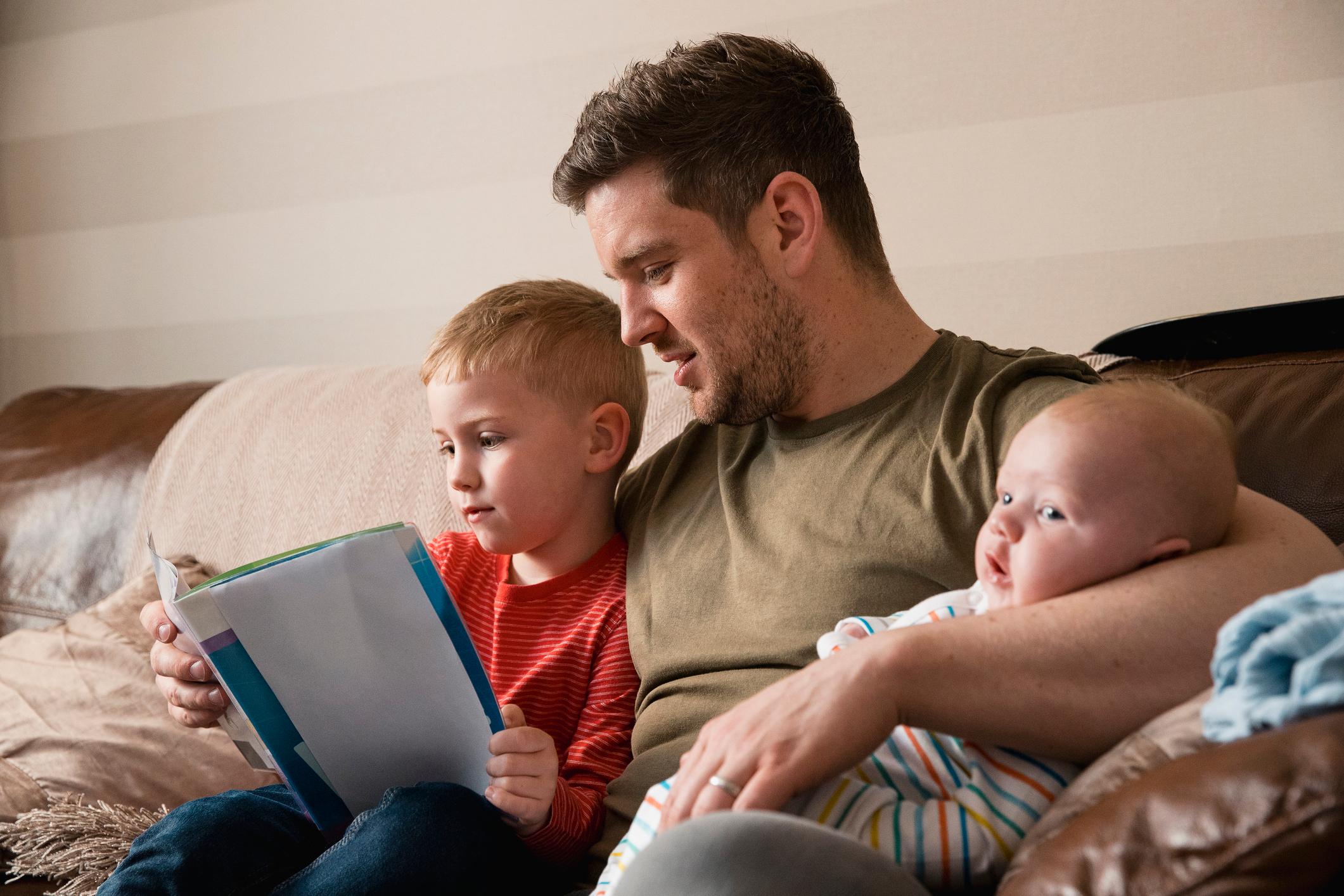 Father holding his newborn baby and reading a book with his older son.