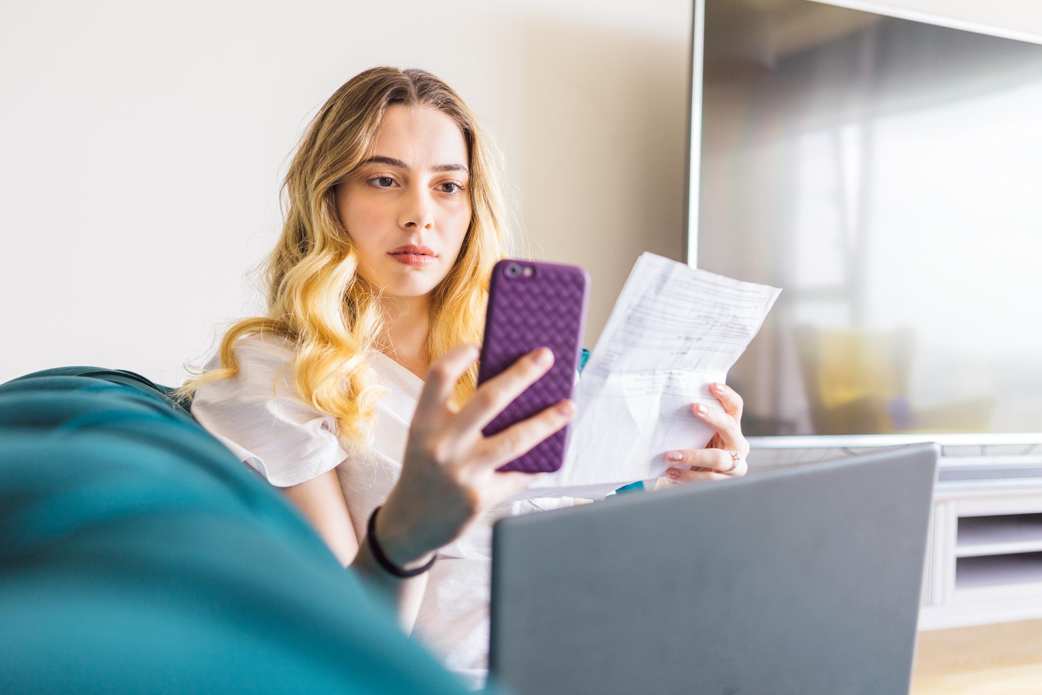 A young woman sat on a sofa with her laptop on lap, looking at her phone, holding a utility bill