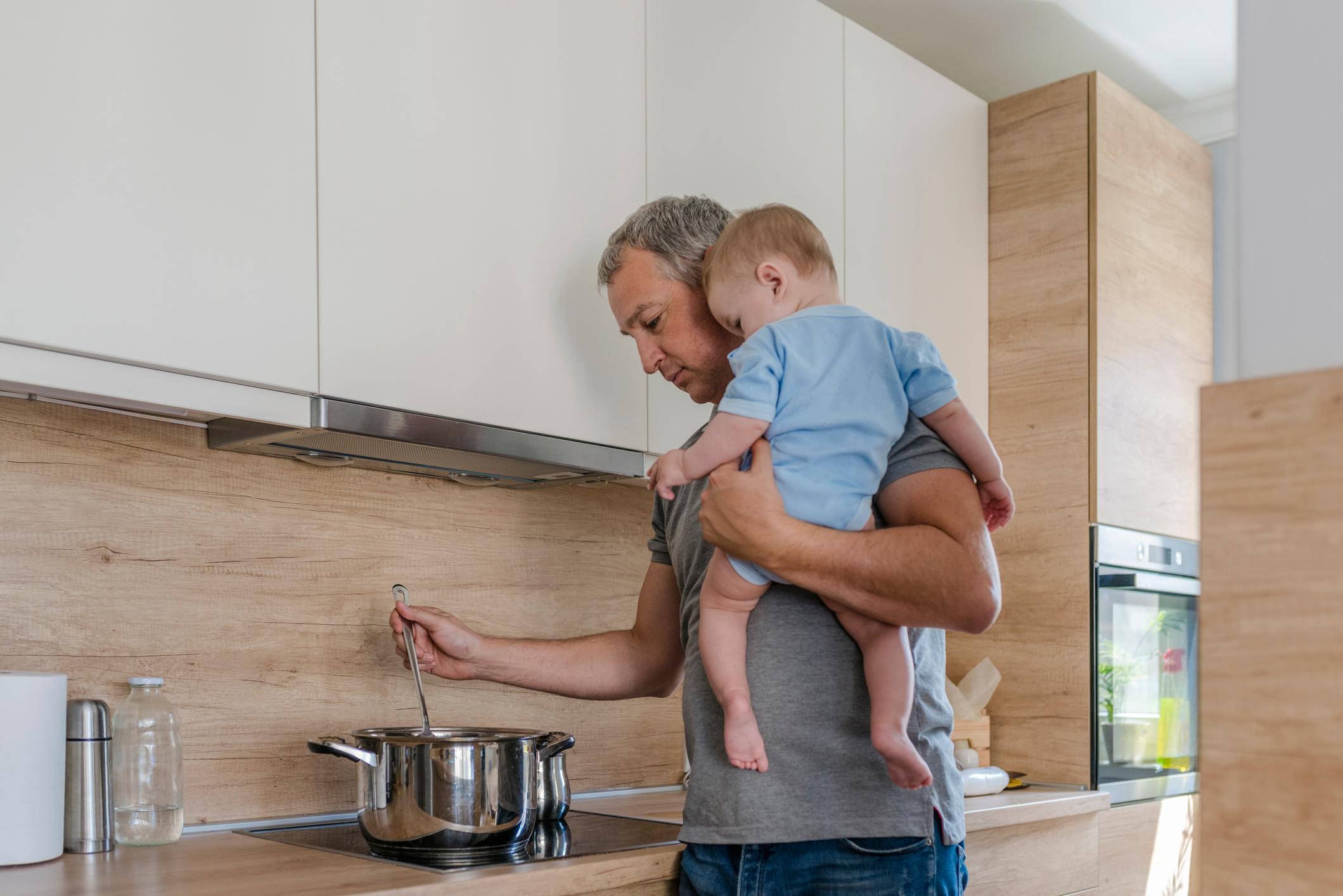 Father in a kitchen holding a child on his shoulder, cooking.