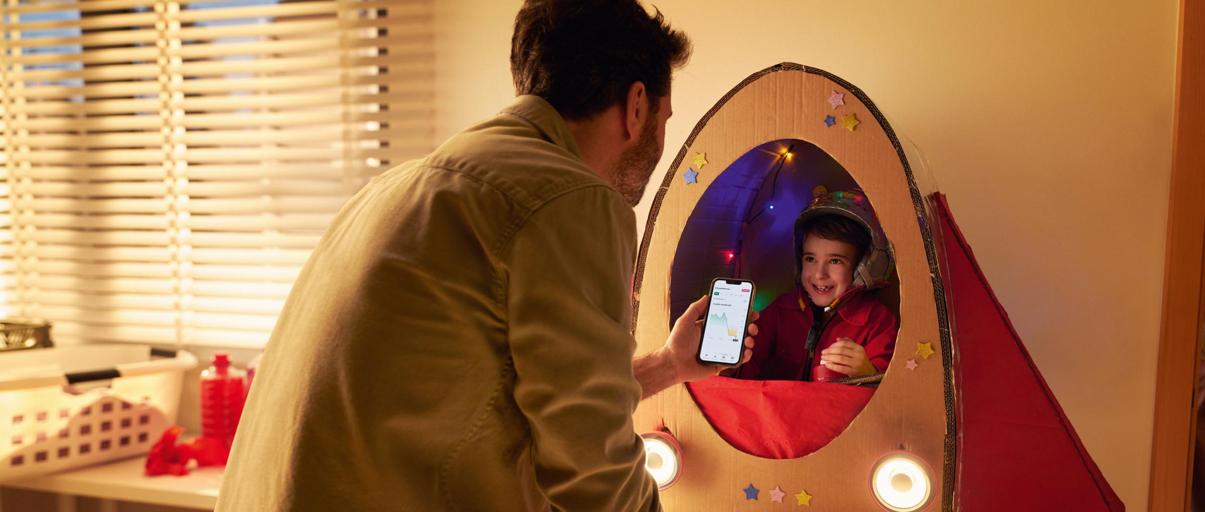Een vader kijkt op de Smart Meter app terwijl zijn zoontje enthousiast in zijn kartonnen ruimteschip bovenop de wasmachine zit.