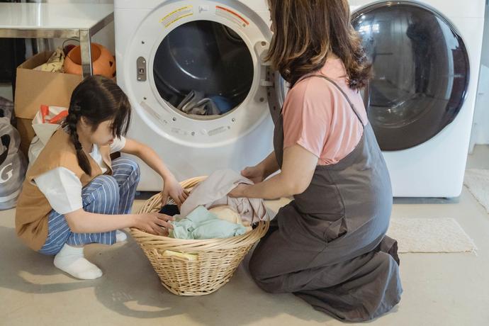 Een moeder en dochter zitten samen bij een wasmachine naast een mand met schone was