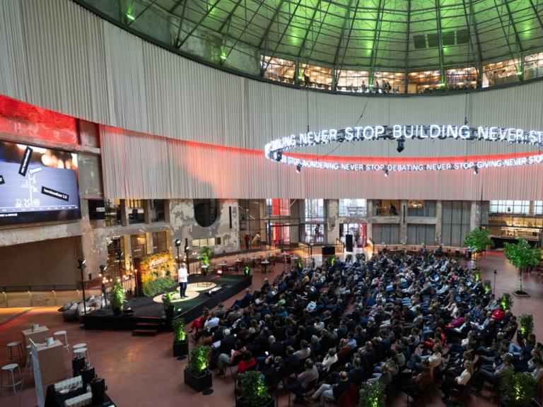 Publiek luistert naar een spreker in de grote zaal van het Wintercircus