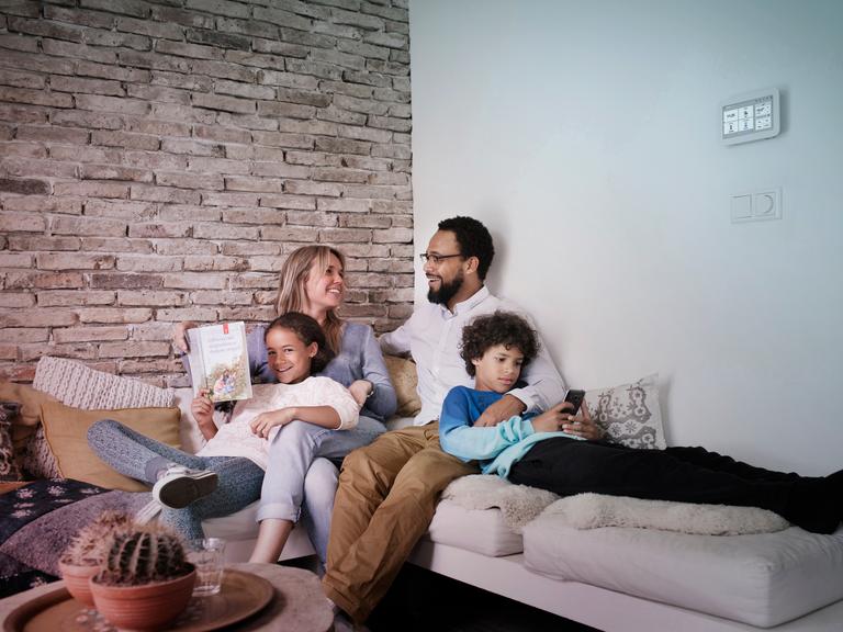 Vader, moeder, dochter en zoon zitten op een hoekbank met een Toon thermostaat aan de muur