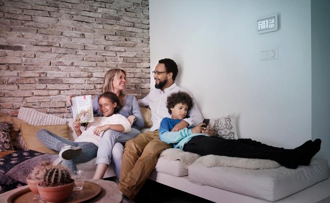 Vader, moeder, dochter en zoon zitten op een hoekbank met een Toon thermostaat aan de muur