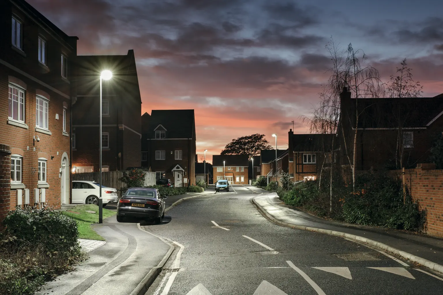 Una tranquilla strada residenziale è illuminata dai lampioni al crepuscolo, con auto parcheggiate lungo la strada. Il cielo si illumina di colori caldi mentre si fa sera.