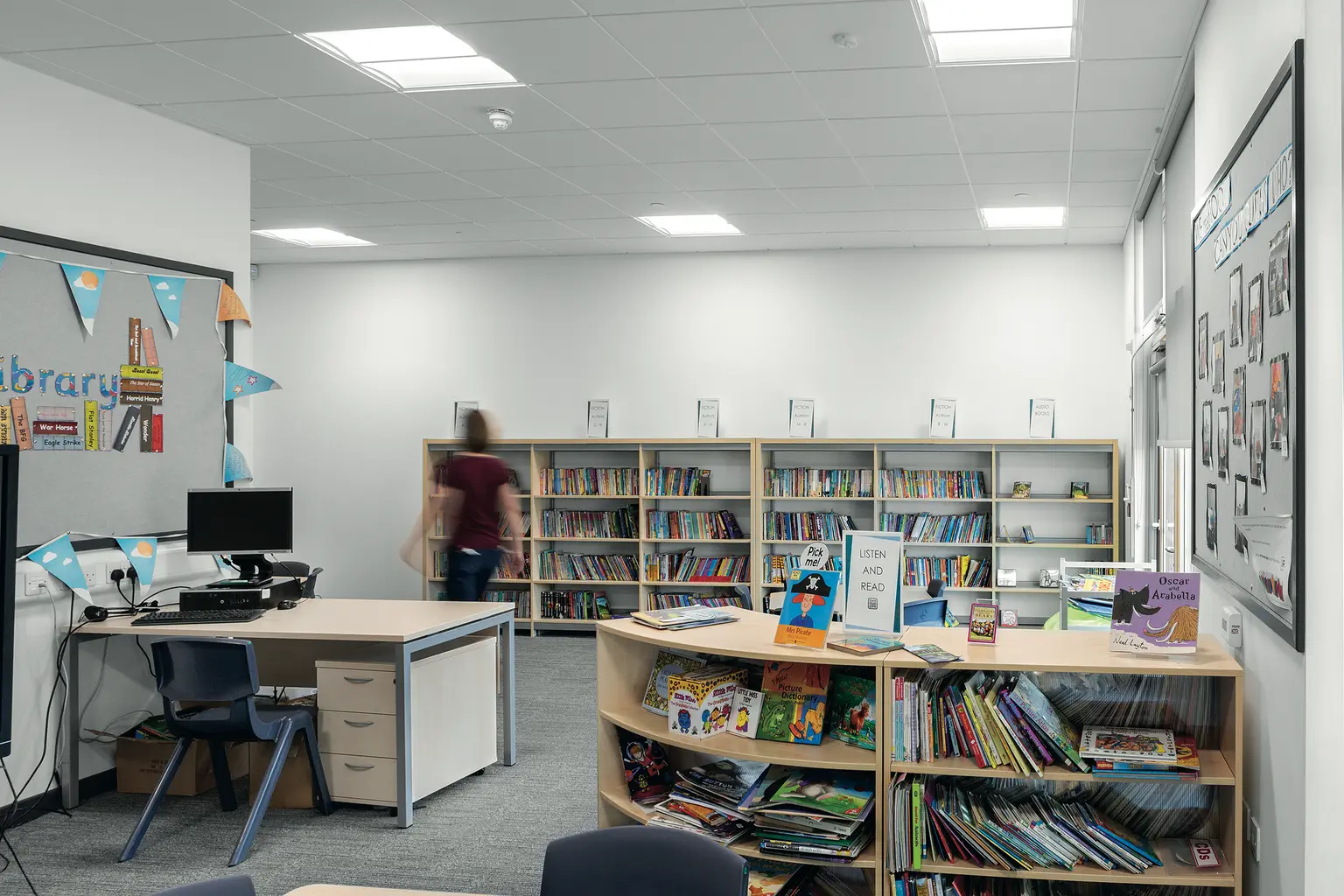 Moderna biblioteca scolastica con scaffali pieni di libri, display curvo e tavolo con computer. Lo spazio è ben illuminato da apparecchi a soffitto e presenta un aspetto sobrio e ordinato, ideale per lo studio.