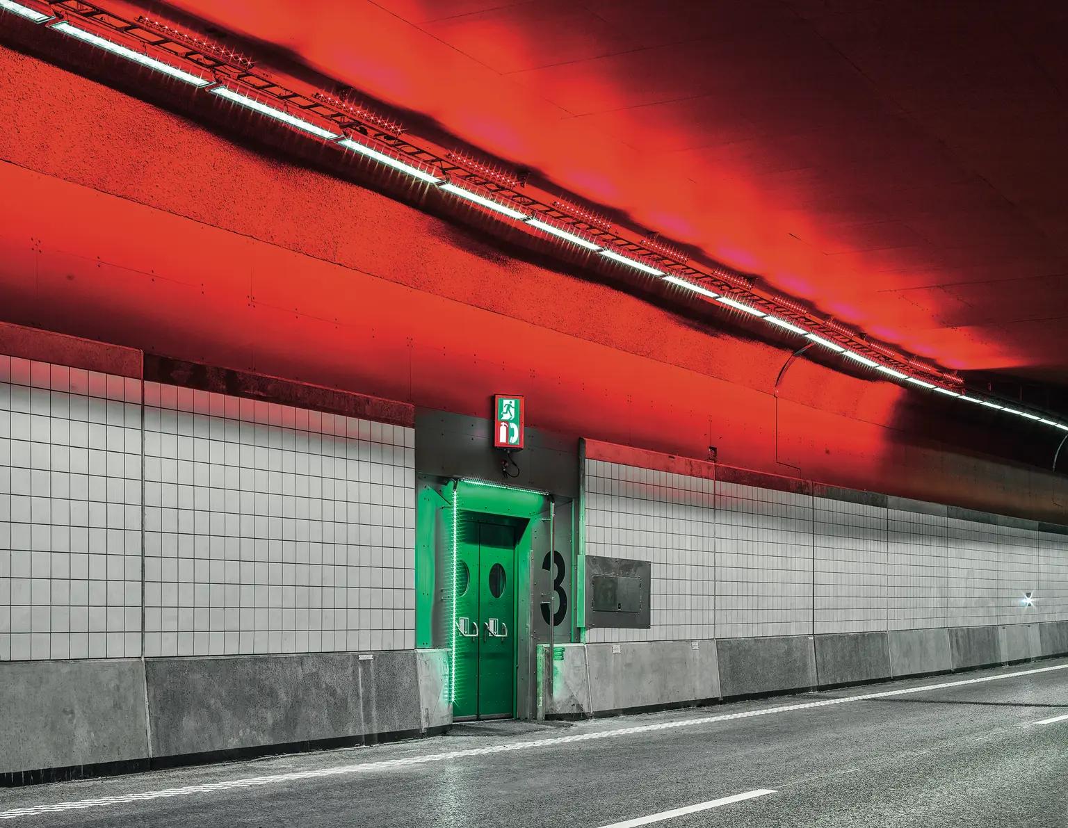 Un tunnel stradale moderno con un suggestivo soffitto rosso illuminato da luci lineari. Lungo le pareti piastrellate si trova una porta di emergenza verde, in audace contrasto con i toni neutri circostanti.