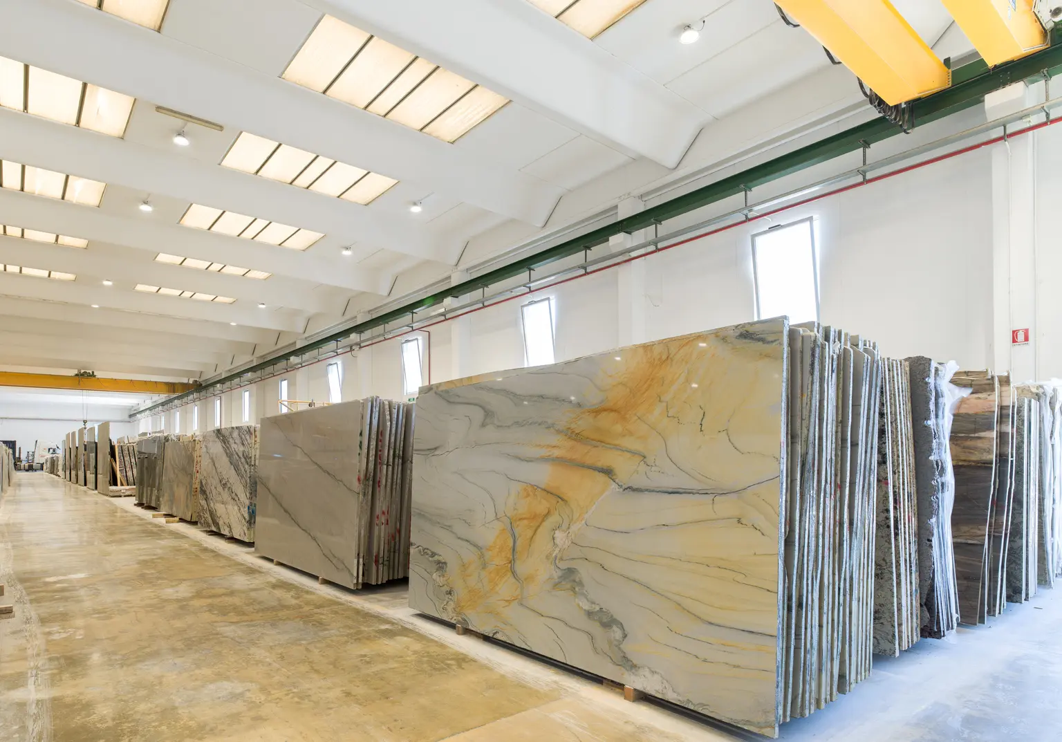 Eine helle Fabrikhalle mit großen Marmorplatten, die fein säuberlich in Reihen angeordnet sind. Der Raum verfügt über hohe Decken, Oberlichter und Kransysteme für einen effizienten Materialtransport.
