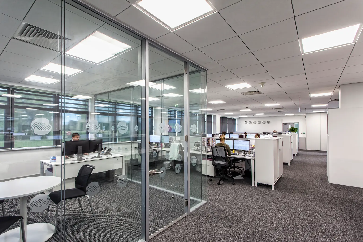 Un ufficio moderno presenta sale riunioni in vetro e file di scrivanie dove i dipendenti lavorano al computer. L'ambiente è spazioso, luminoso e ordinato.