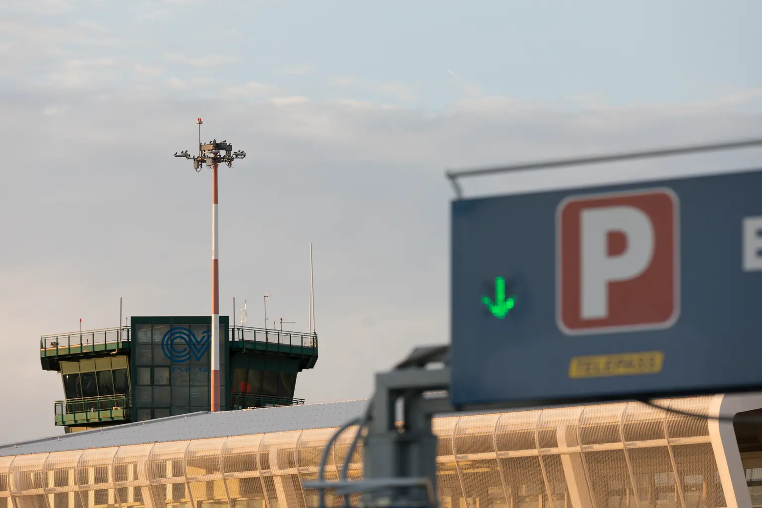 Torre di controllo dell'aeroporto ed edificio del terminal al tramonto, con un cartello di parcheggio e una freccia verde in primo piano. La scena è illuminata dolcemente con un cielo limpido sopra.