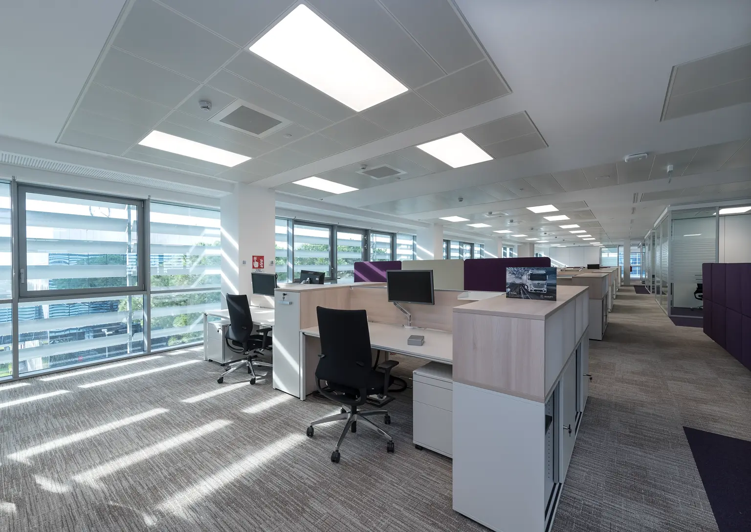 Un ufficio moderno con un layout a pianta aperta presenta lunghe file di scrivanie, divisori viola e una sala riunioni con pareti in vetro e arredamento minimal. Ambiente luminoso e pulito illuminato da pannelli LED.