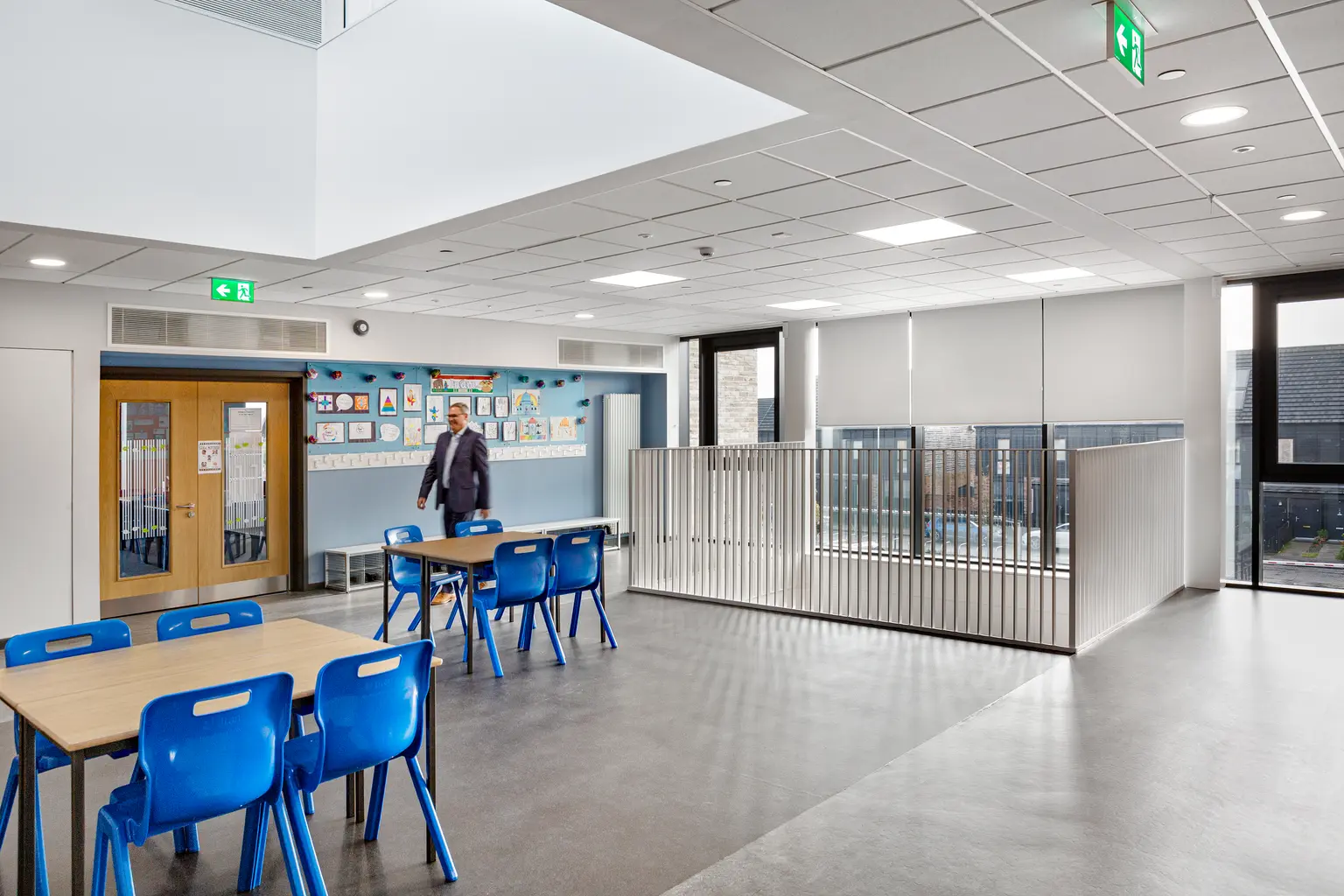 Ambiente scolastico luminoso e spazioso con sedie azzurre, tavoli in legno e ampie finestre che permettono alla luce naturale di entrare. Include bacheche alle pareti e una moderna illuminazione a soffitto che conferisce allo spazio un aspetto sobrio e ordinato.