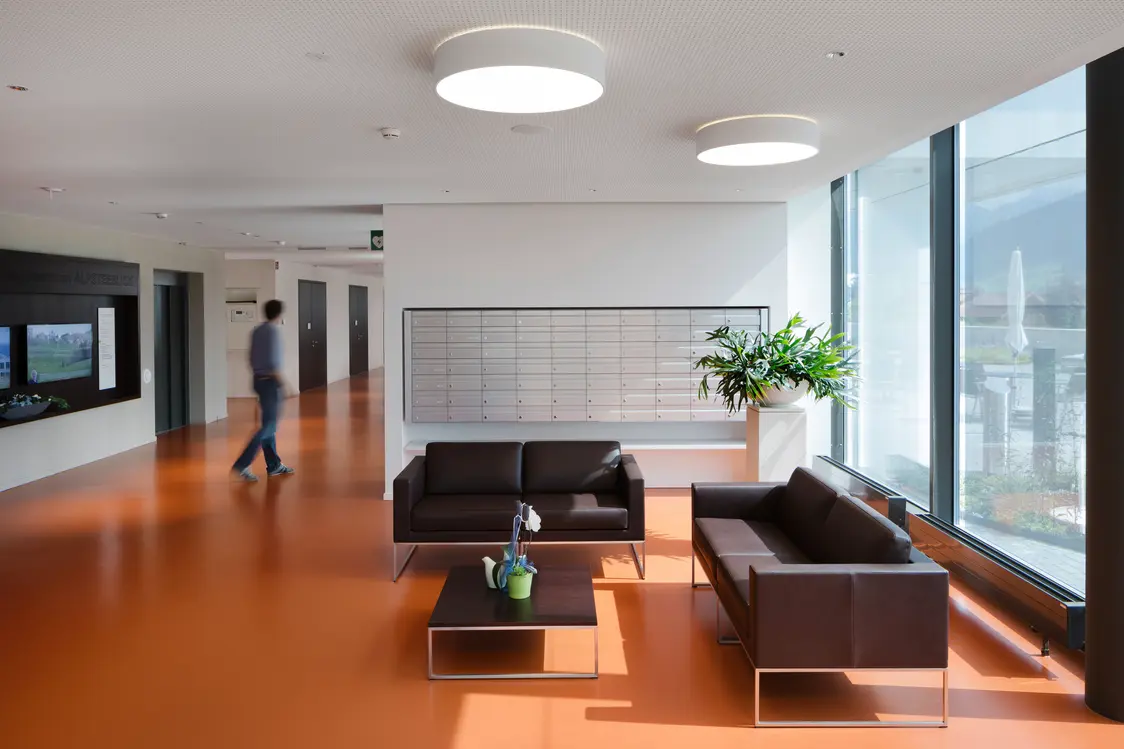 Ein moderner, heller Empfangsbereich mit braunen Ledersofas, einem Couchtisch und großen Fenstern, durch die Tageslicht einfällt. Eine Person geht im Hintergrund durch den Raum, der mit minimalistischen Lampen und einer großen Grünpflanze dekoriert ist.