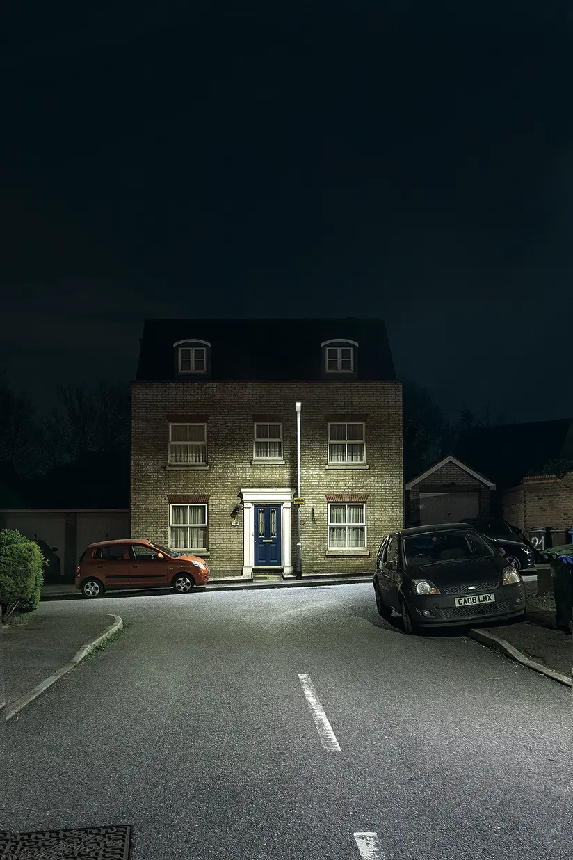 Ein Backsteinhaus steht am Ende einer ruhigen Wohnstraße bei Nacht, beleuchtet von einer einzelnen Straßenleuchte. Vor dem Haus sind zwei geparkte Autos sichtbar, die Szene wirkt ruhig und leer.