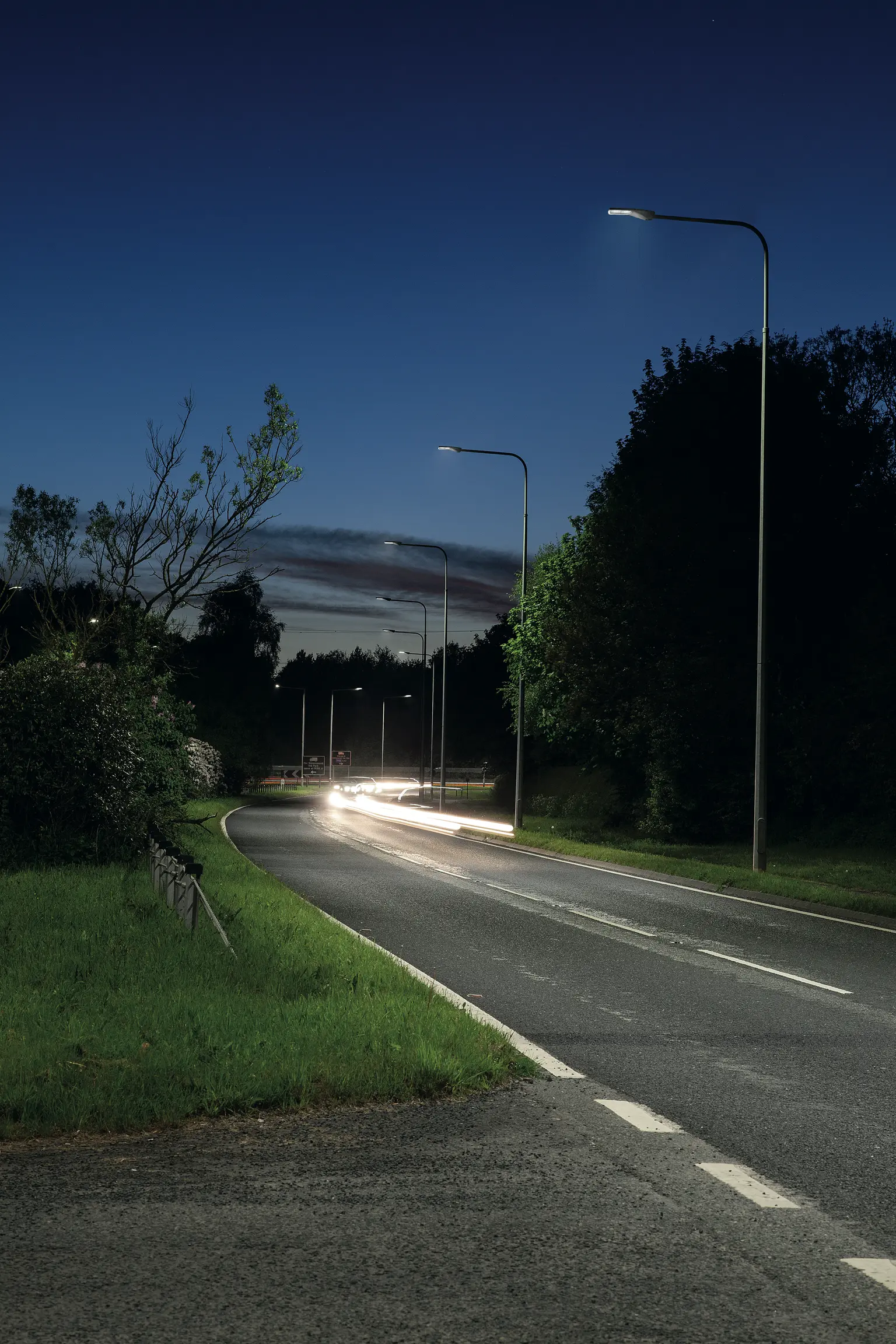 Eine kurvenreiche Vorortstraße in der Abenddämmerung, beleuchtet von modernen Straßenlaternen und umgeben von Bäumen und Grünflächen. Die Lichtstrahlen der vorbeifahrenden Autos erzeugen einen dynamischen Effekt unter dem dunkelblauen Abendhimmel.