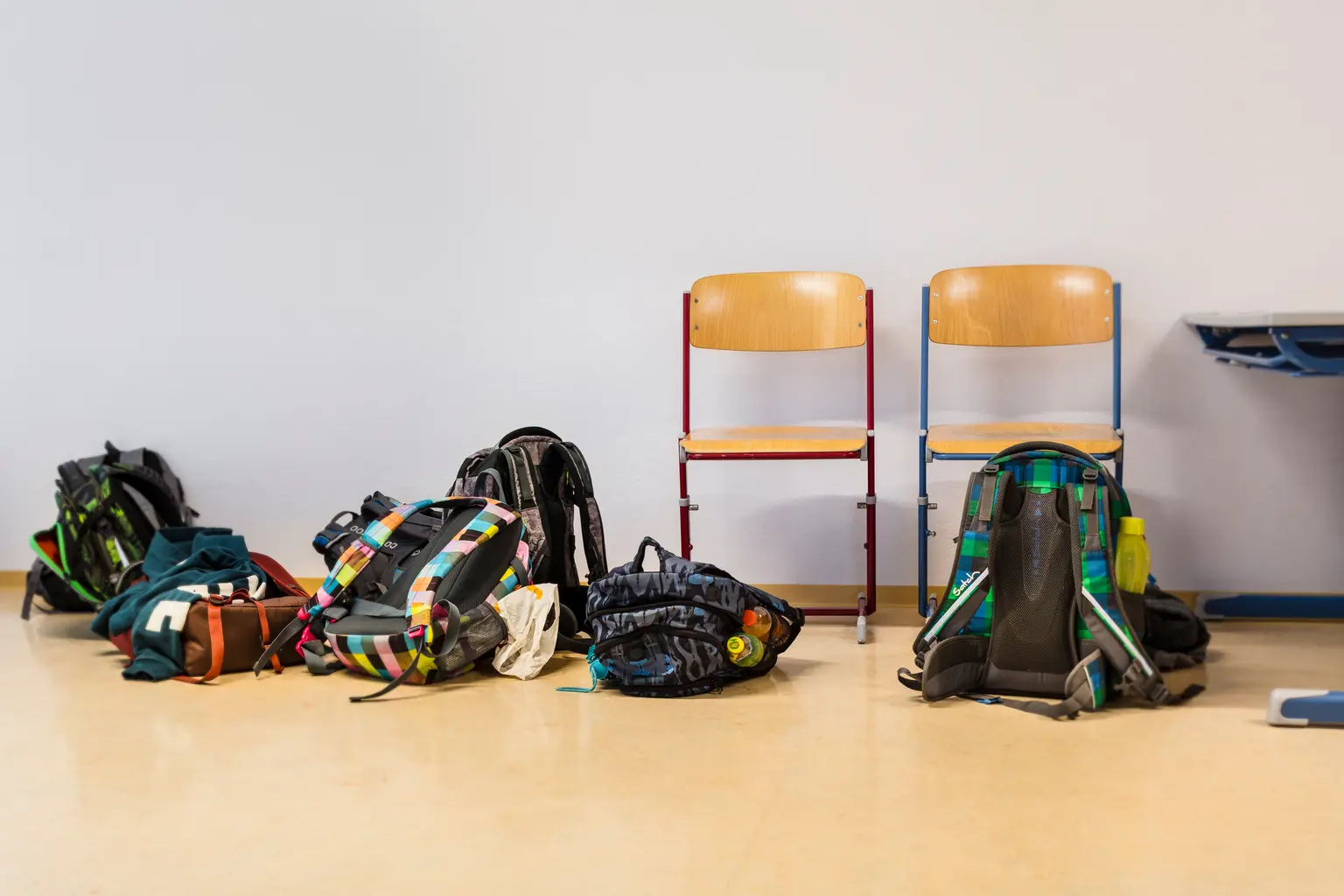 Diversi zaini sono posizionati a terra accanto a due sedie di legno con una parete bianca sullo sfondo. La scena è ambientata in un’aula con pavimento chiaro.