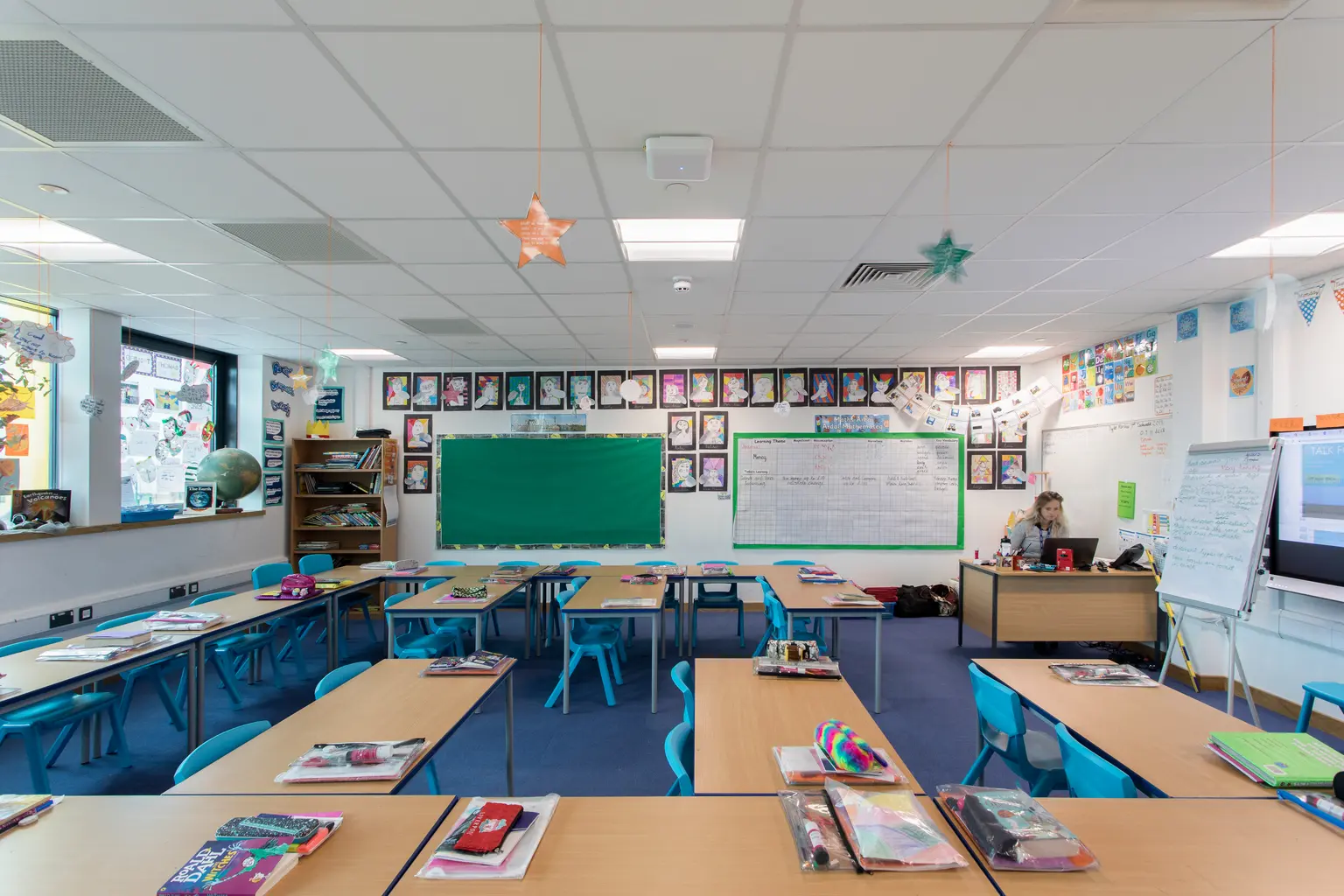 Un'aula luminosa e ordinata con banchi di legno, sedie azzurre, una lavagna tradizionale verde e una lavagna bianca. Le pareti sono decorate con opere d'arte degli studenti e il locale è ben illuminato da luci a soffitto e luce naturale che penetra dalle grandi finestre.