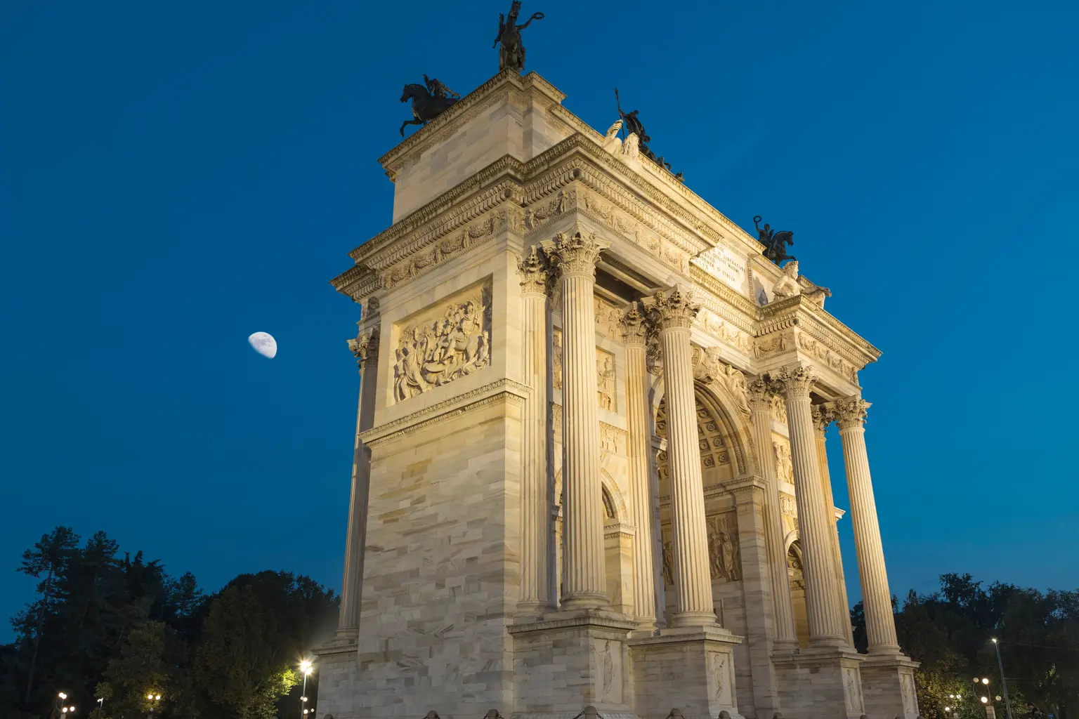 L'Arco della Pace è visto da un angolo al crepuscolo, con i dettagli architettonici messi in risalto dall'illuminazione e dalla luna nel cielo.