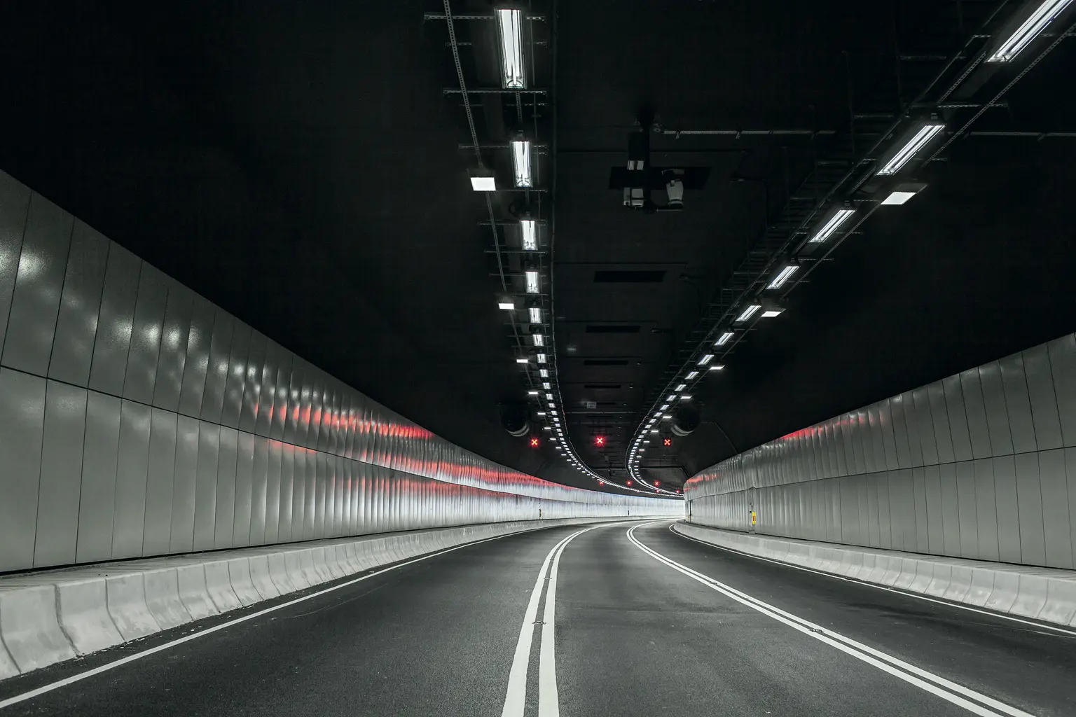 Un tunnel moderno e vuoto con intense luci sopraelevate e segnaletica orizzontale bianca che curva dolcemente verso sinistra. Le pareti del tunnel, lisce e riflettenti, conferiscono un aspetto pulito e simmetrico.