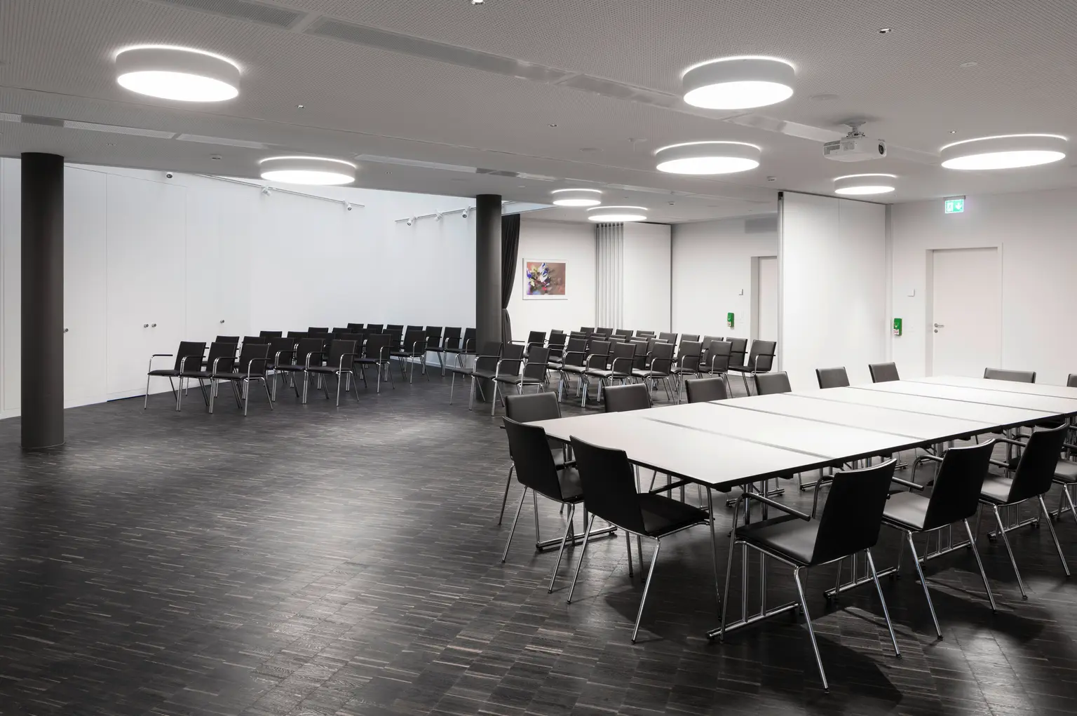 Ein leerer Konferenzraum mit Tischen und Stühlen, vorbereitet für eine Veranstaltung. Das helle Design und die moderne Beleuchtung schaffen eine professionelle Atmosphäre.