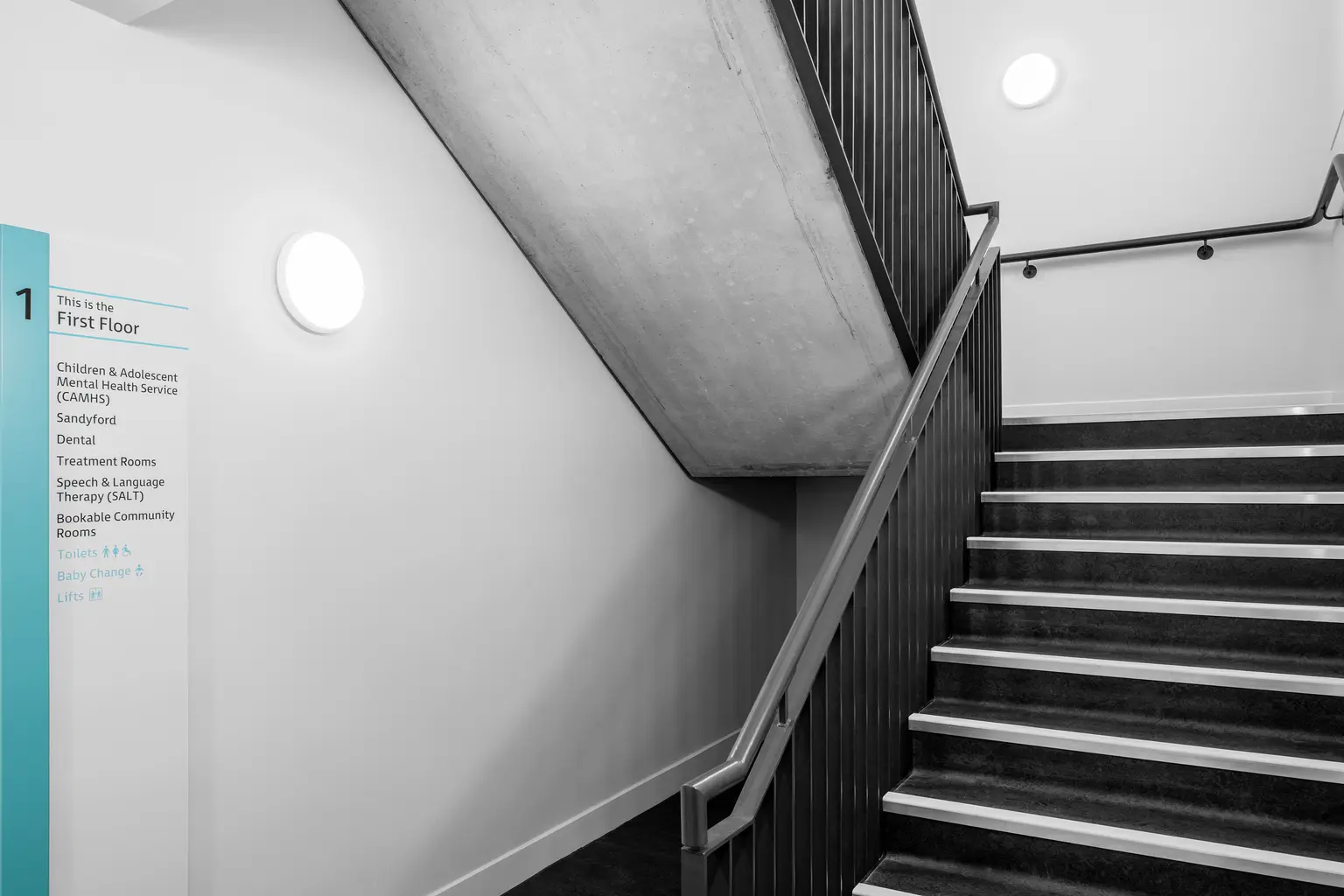 Un'area della scala illuminata da luci LED circolari montate a parete. Il design minimalista presenta ringhiere nere, elementi in cemento e segnaletica direzionale chiara che conferiscono un aspetto pulito e pratico.