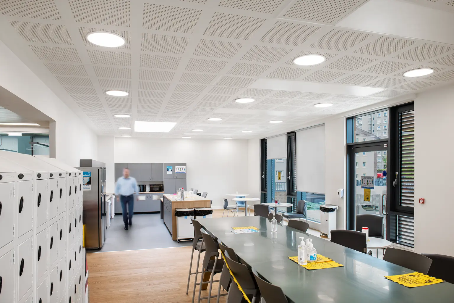 Una caffetteria moderna con luci LED luminose integrate in un soffitto a griglia. È presente un lungo tavolo da pranzo, armadietti e grandi finestre che forniscono luce naturale, creando un ambiente aperto e accogliente.