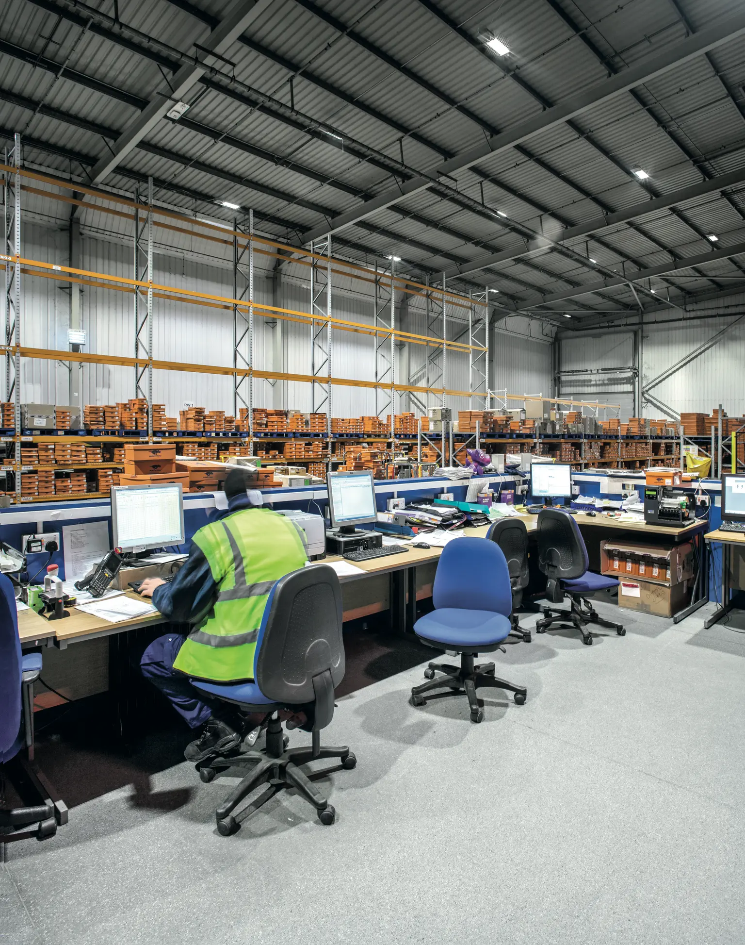Office area within a warehouse, several workstations with computers and a worker in a safety vest; the lighting solution with ceiling-mounted LED lights ensures a well-lit and productive workspace.