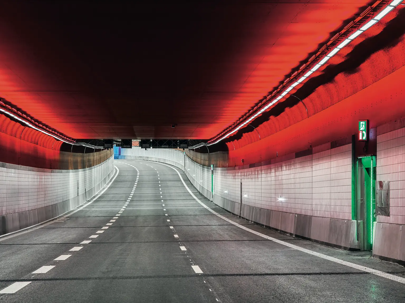 Un tunnel stradale moderno con una corsia leggermente curva, illuminato da luci rosse a soffitto e illuminazione bianca a parete. Le pareti sono rivestite di piastrelle e sulla destra è visibile una porta verde di uscita di emergenza.