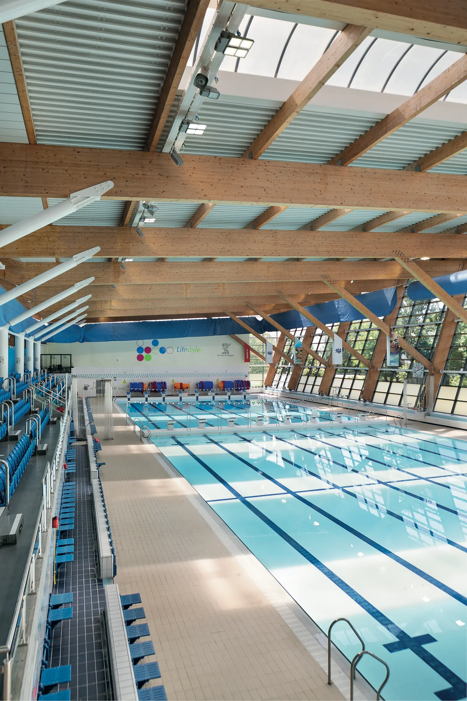 L'interno di un moderno impianto di piscina coperta con corsie multiple, circondata da posti a sedere color azzurro. Travi in legno e grandi finestre consentono alla luce naturale di riempire lo spazio, creando un'atmosfera luminosa e aperta. La qualità dell'illuminazione migliora la sicurezza, la visibilità e l'esperienza complessiva per atleti e visitatori.